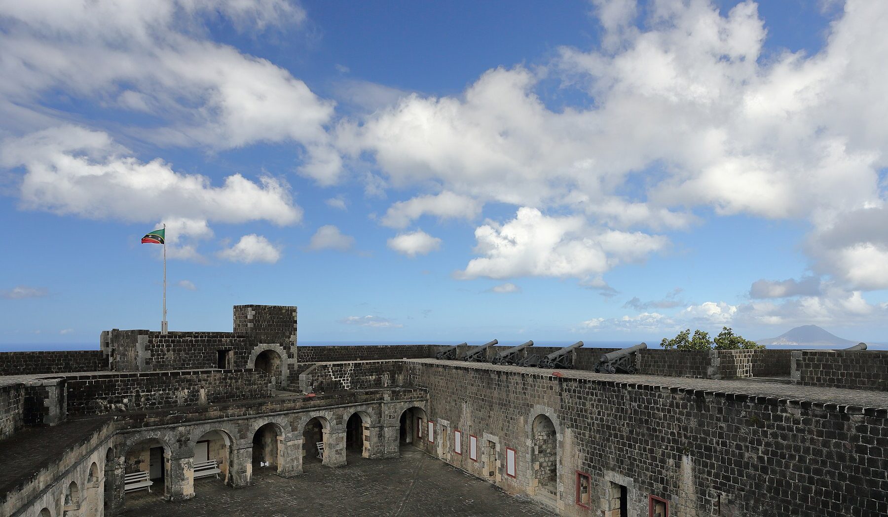 Stone courtyard and cannon-lined walls of Fort George Citadel at Brimstone Hill with the Saint Kitts and Nevis flag and the sea beyond