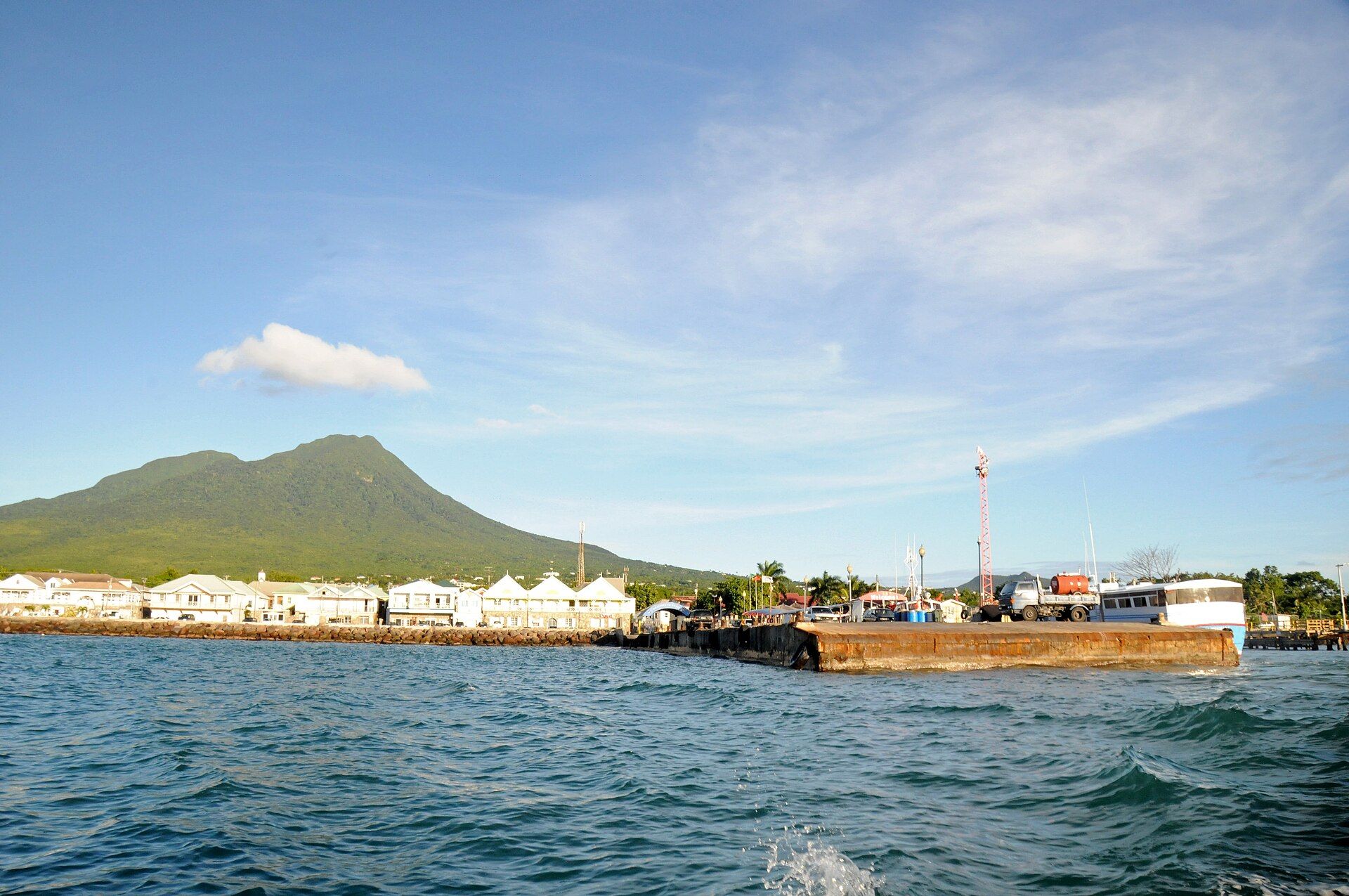 Green volcanic peak rising behind the waterfront buildings and a cargo pier of Charlestown, Nevis, seen from the sea
