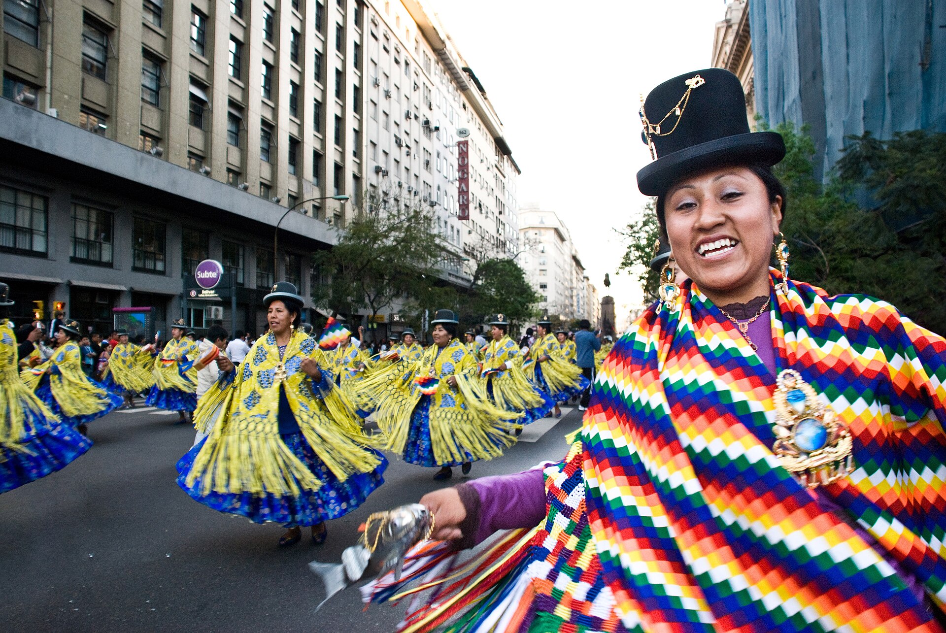 Women in bowler hats and bright layered skirts and shawls dance in a street parade between office buildings in central Buenos Aires