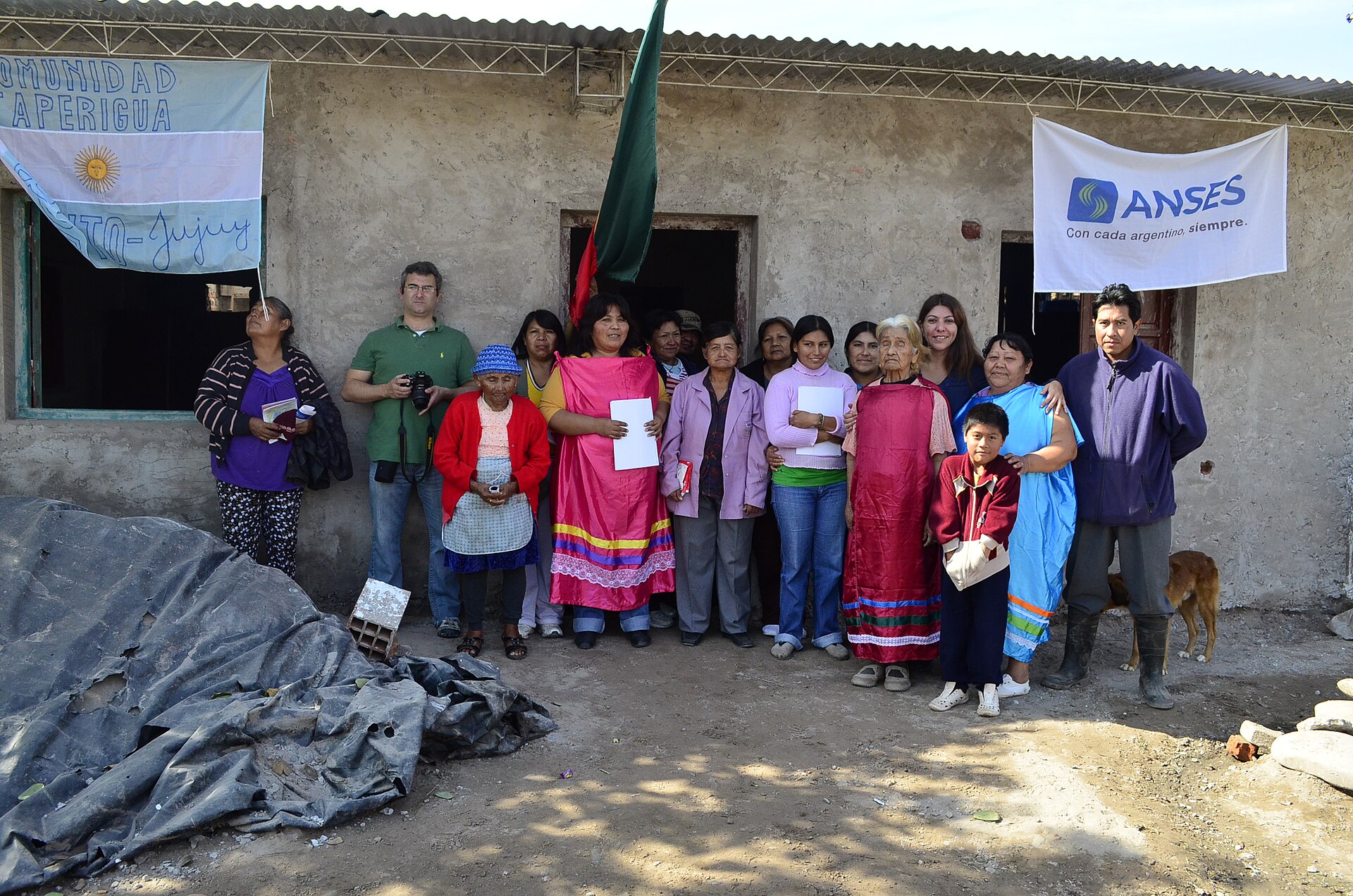 A group of adults and a child stand outside a simple house hung with an Argentine flag reading Comunidad Taperigua and an ANSES banner