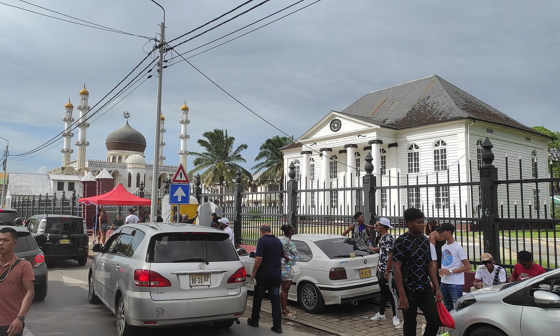 A white mosque with a dark dome and minarets on the left, a white wooden synagogue with a pediment and clock on the right, and parked cars and pedestrians in the foreground