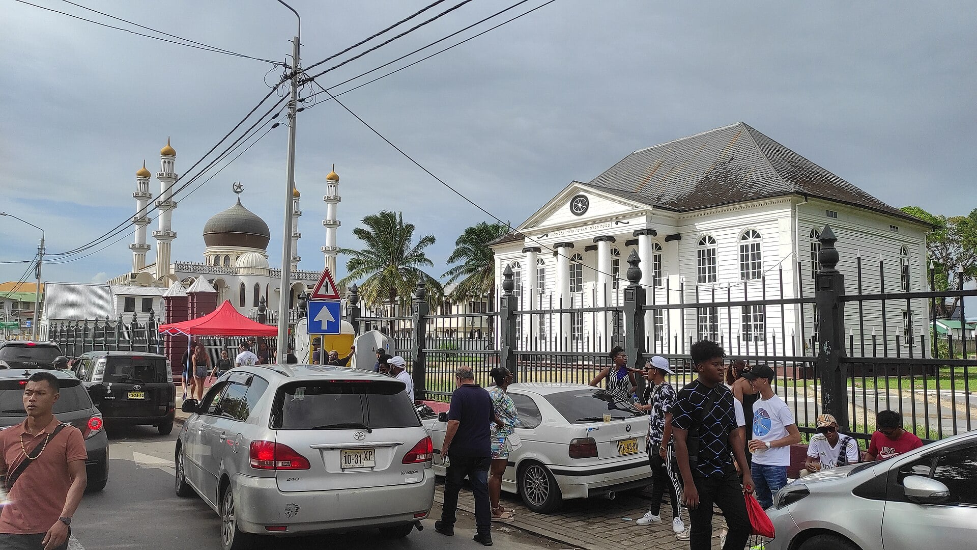 A white mosque with a dark dome and minarets on the left, a white wooden synagogue with a pediment and clock on the right, and parked cars and pedestrians in the foreground