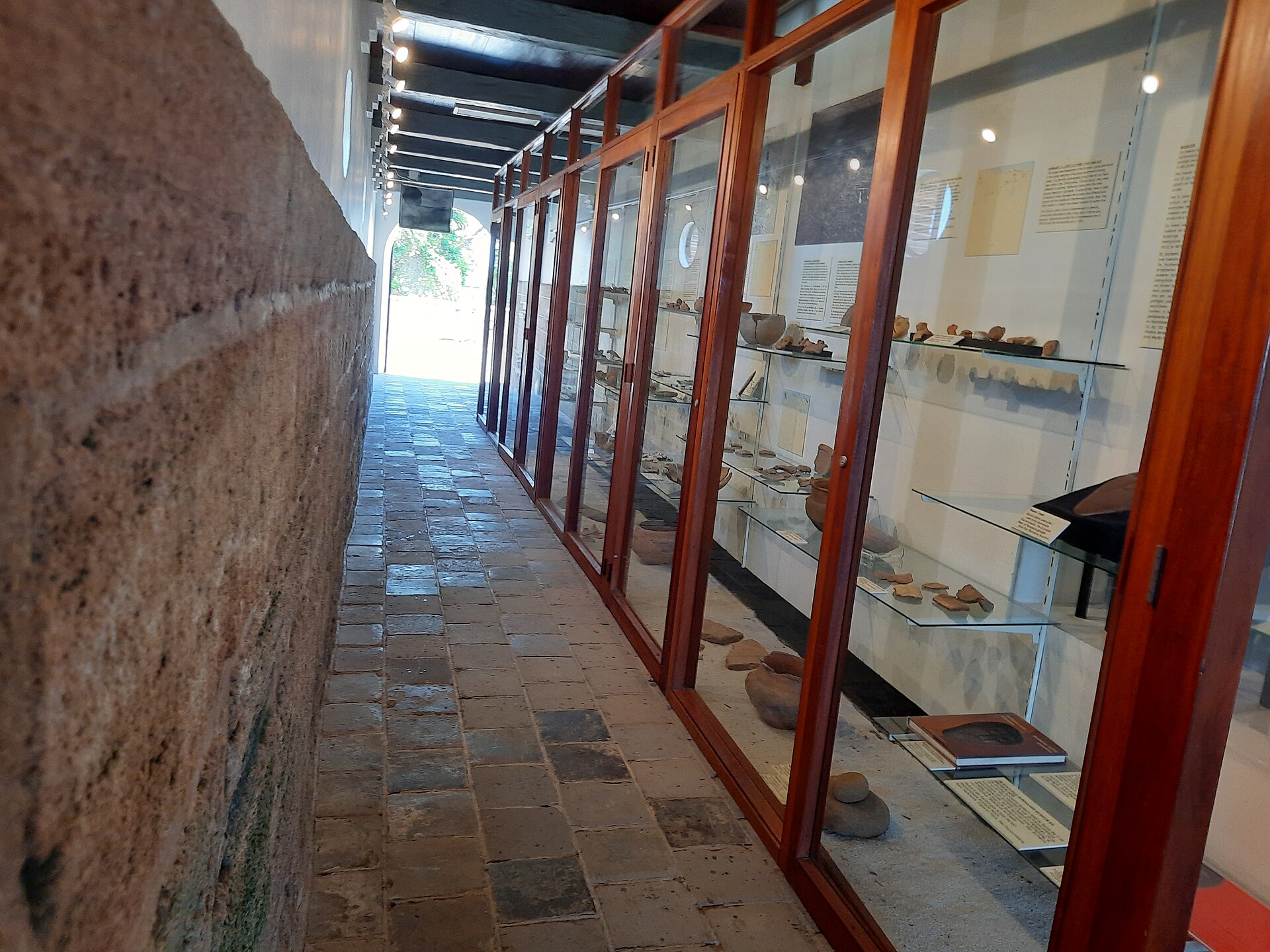 A stone-floored corridor in Fort Zeelandia lined with glass display cases of pottery fragments, with a brick wall on the left