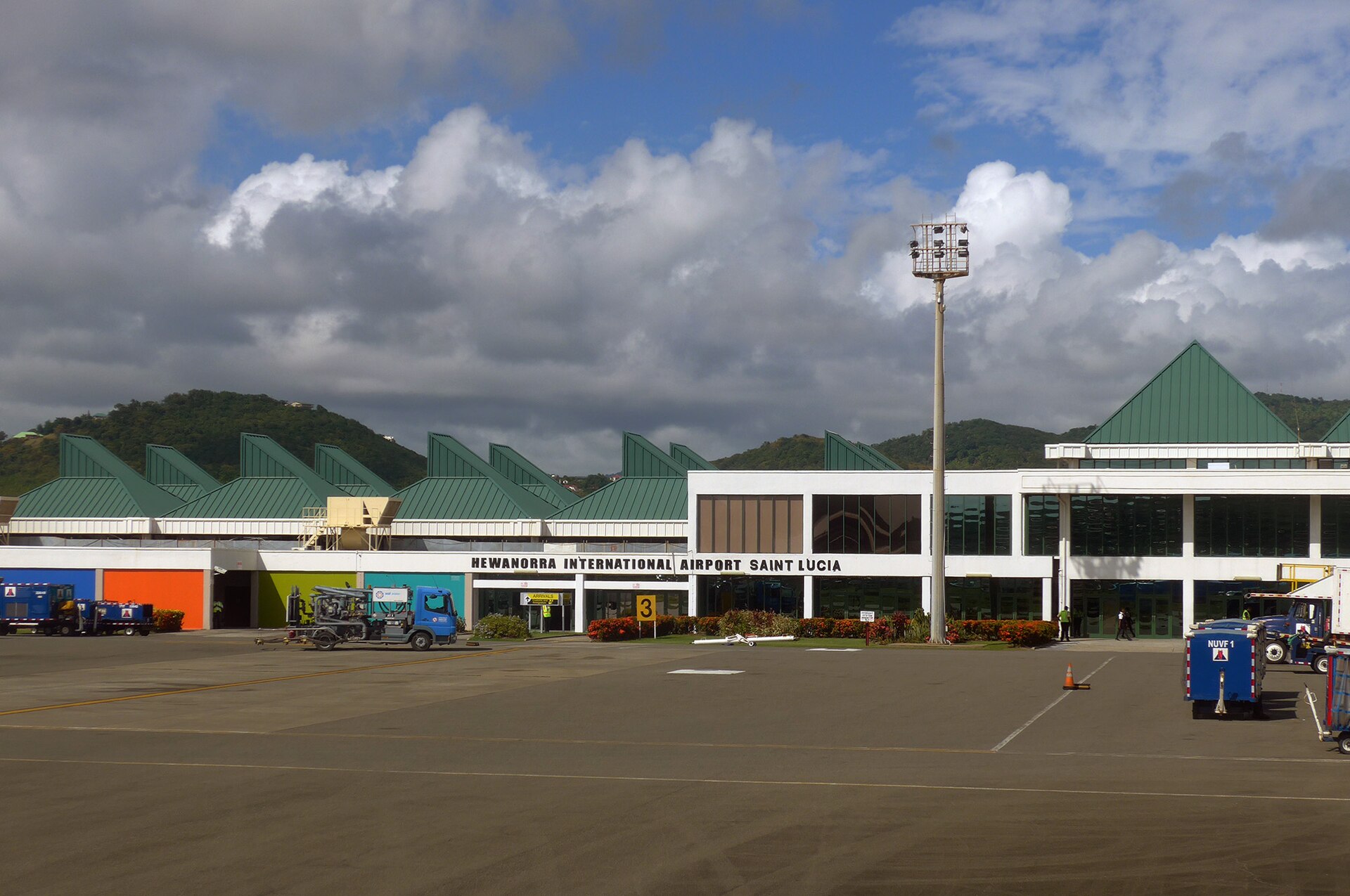 Green-roofed terminal building of Hewanorra International Airport with an apron, service trucks and wooded hills behind