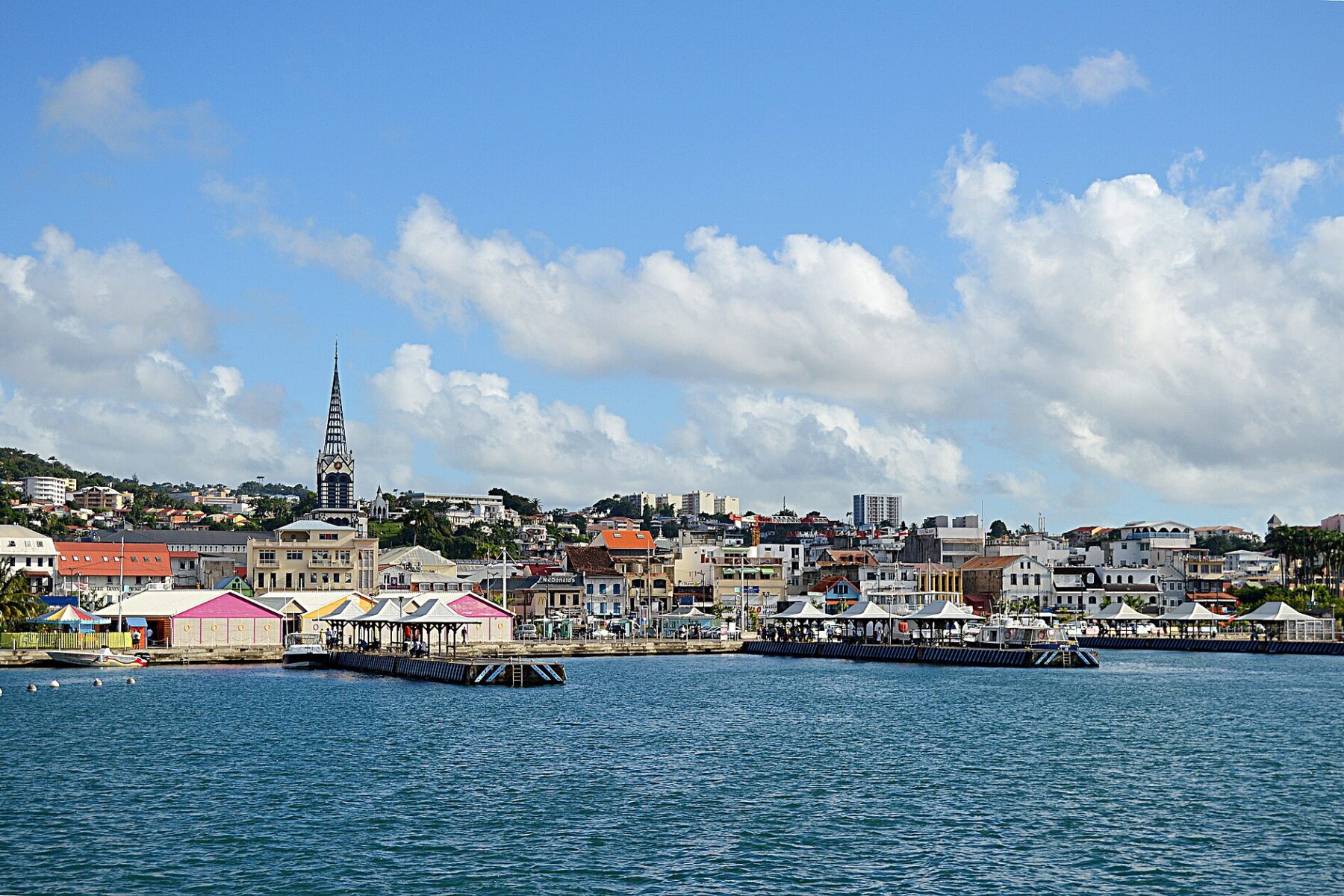 Waterfront of Fort-de-France with a church spire, market pavilions and colourful buildings climbing a hill under a blue sky