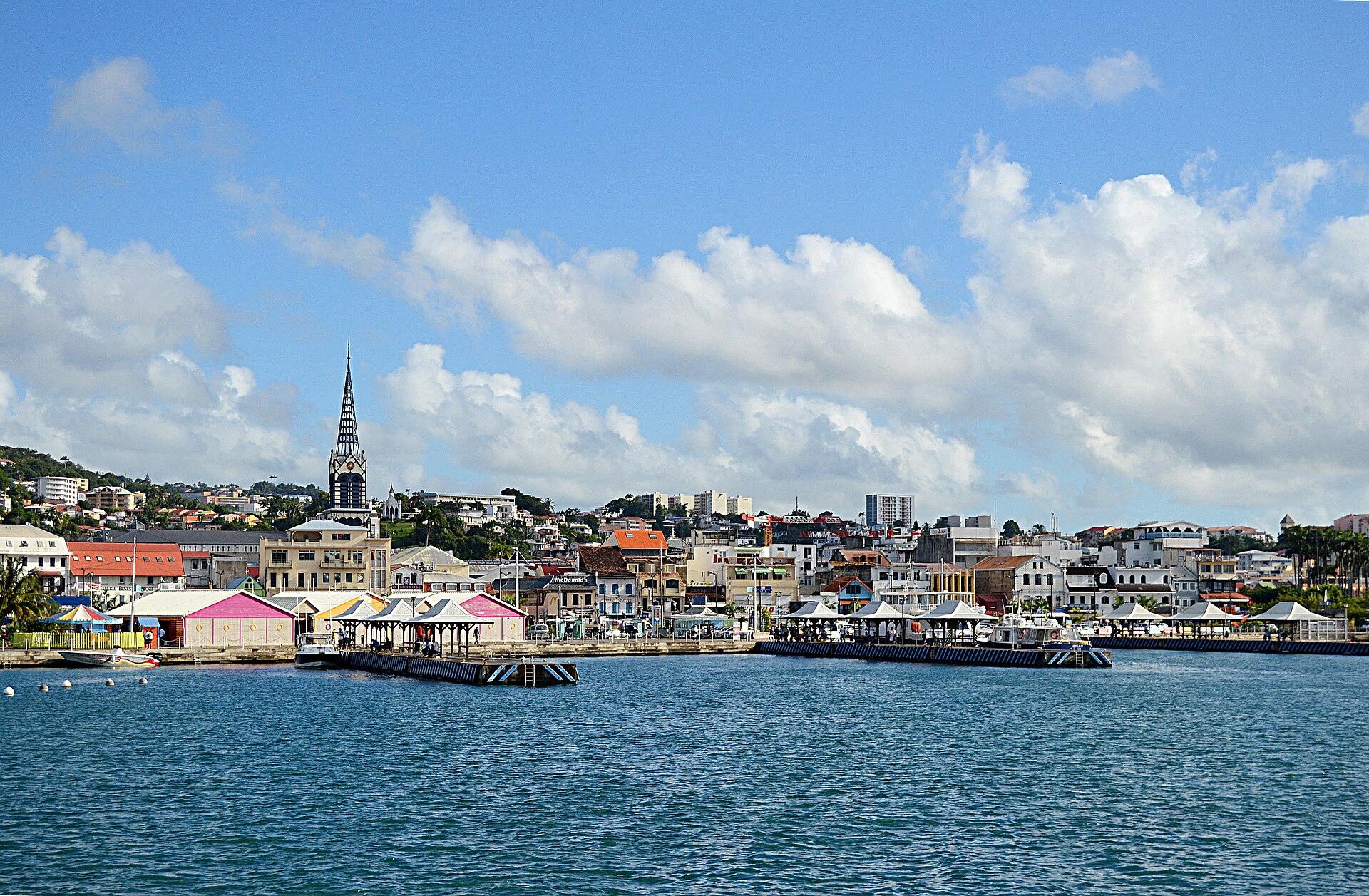 Waterfront of Fort-de-France with a church spire, market pavilions and colourful buildings climbing a hill under a blue sky