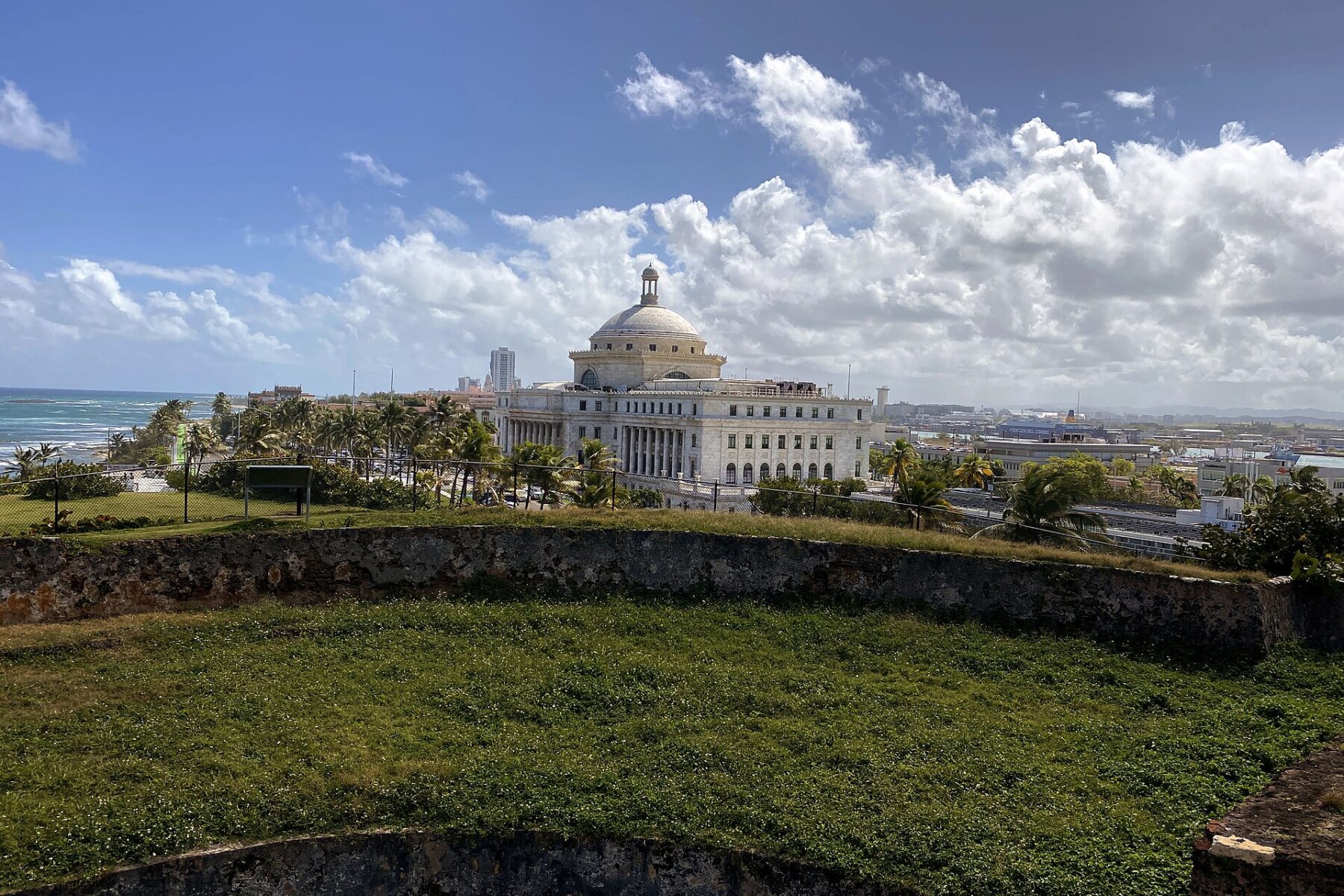 The white domed Capitol of Puerto Rico rises behind palm trees and the grassy stone walls of San Cristóbal Fortress, with the Atlantic Ocean to the left under a blue sky