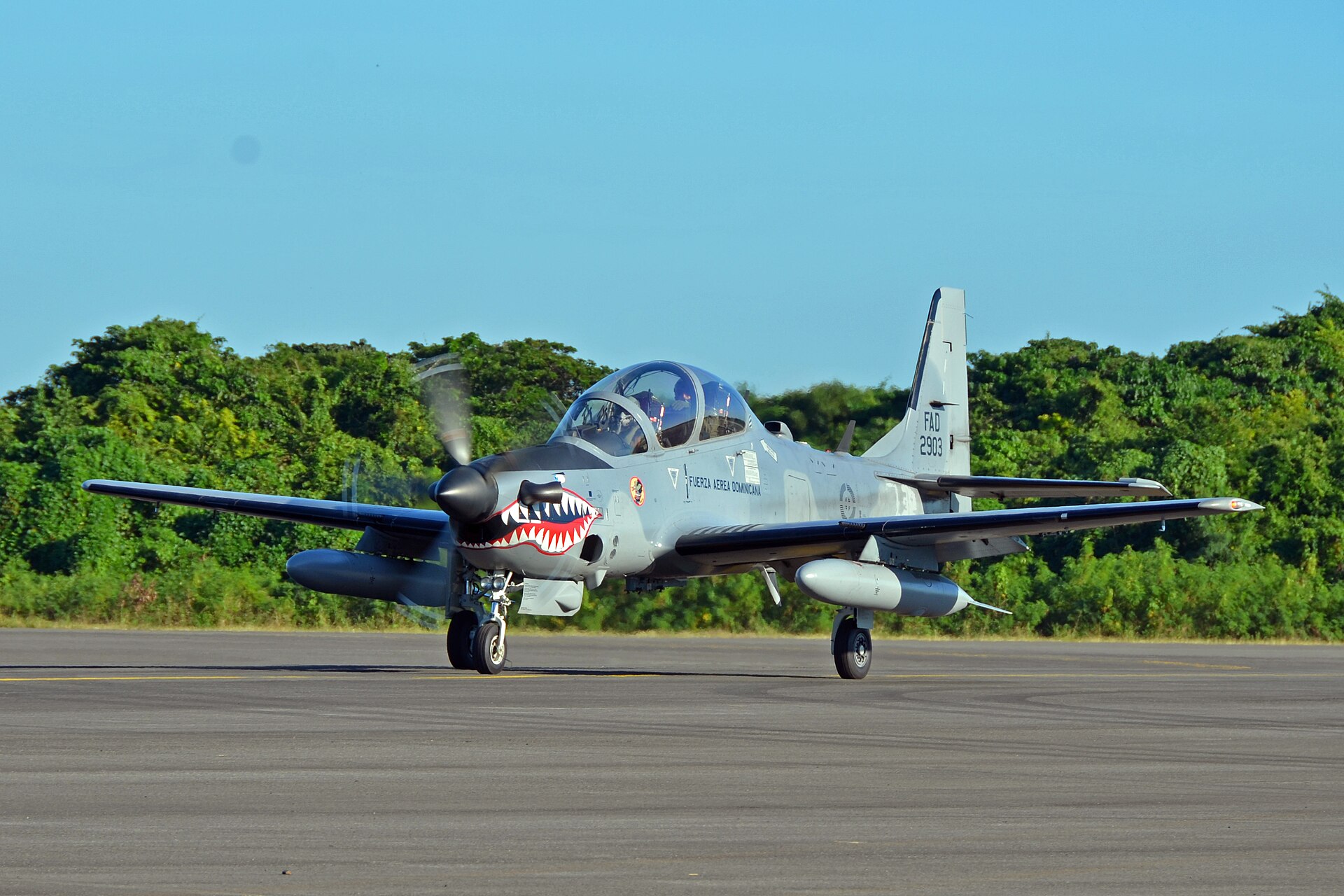 A grey Dominican air force A-29 Super Tucano propeller aircraft with a shark-mouth nose taxis on a runway in front of green trees under a clear blue sky
