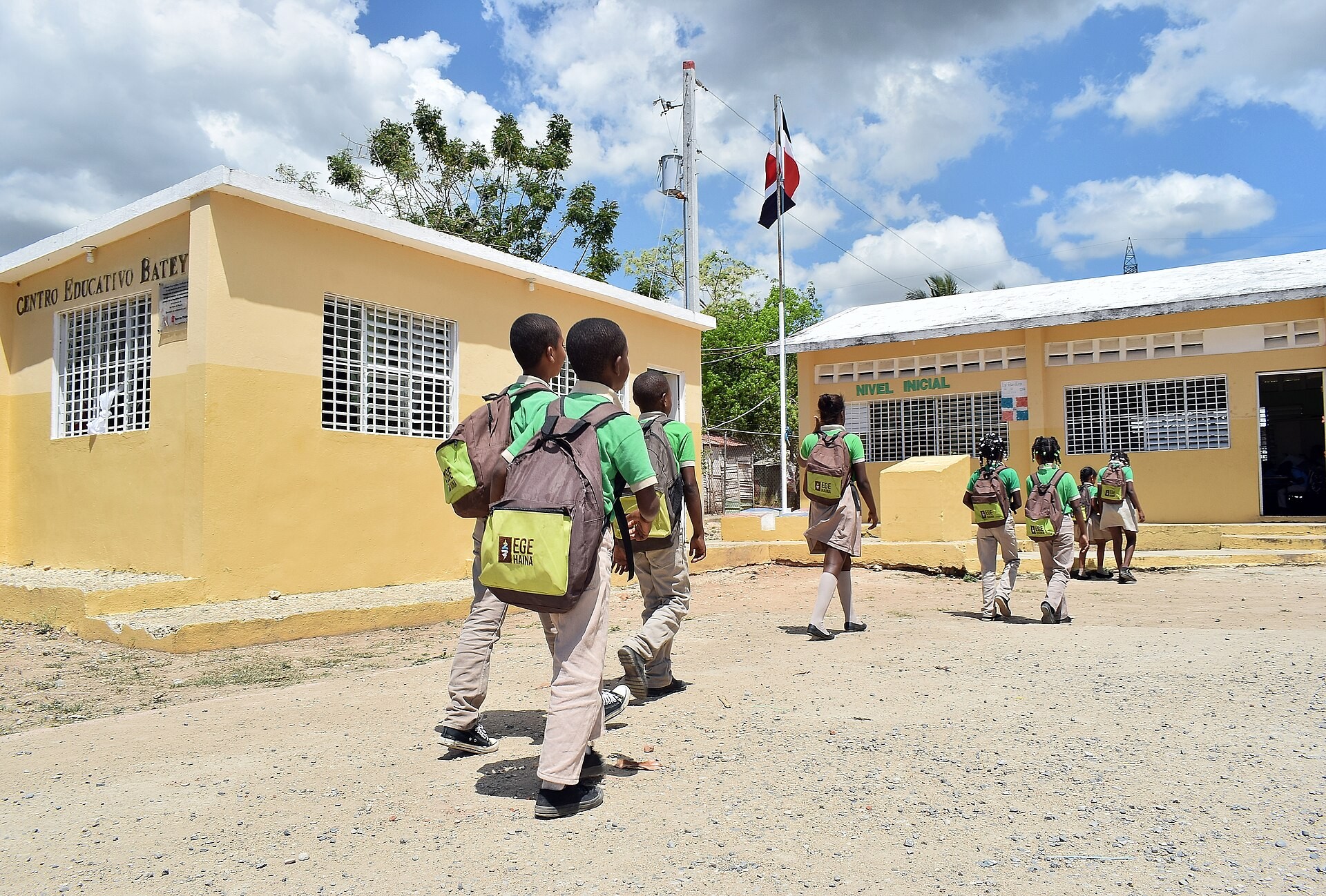 Schoolchildren in green shirts and backpacks walk toward yellow single-storey school buildings with a Dominican flag on a pole