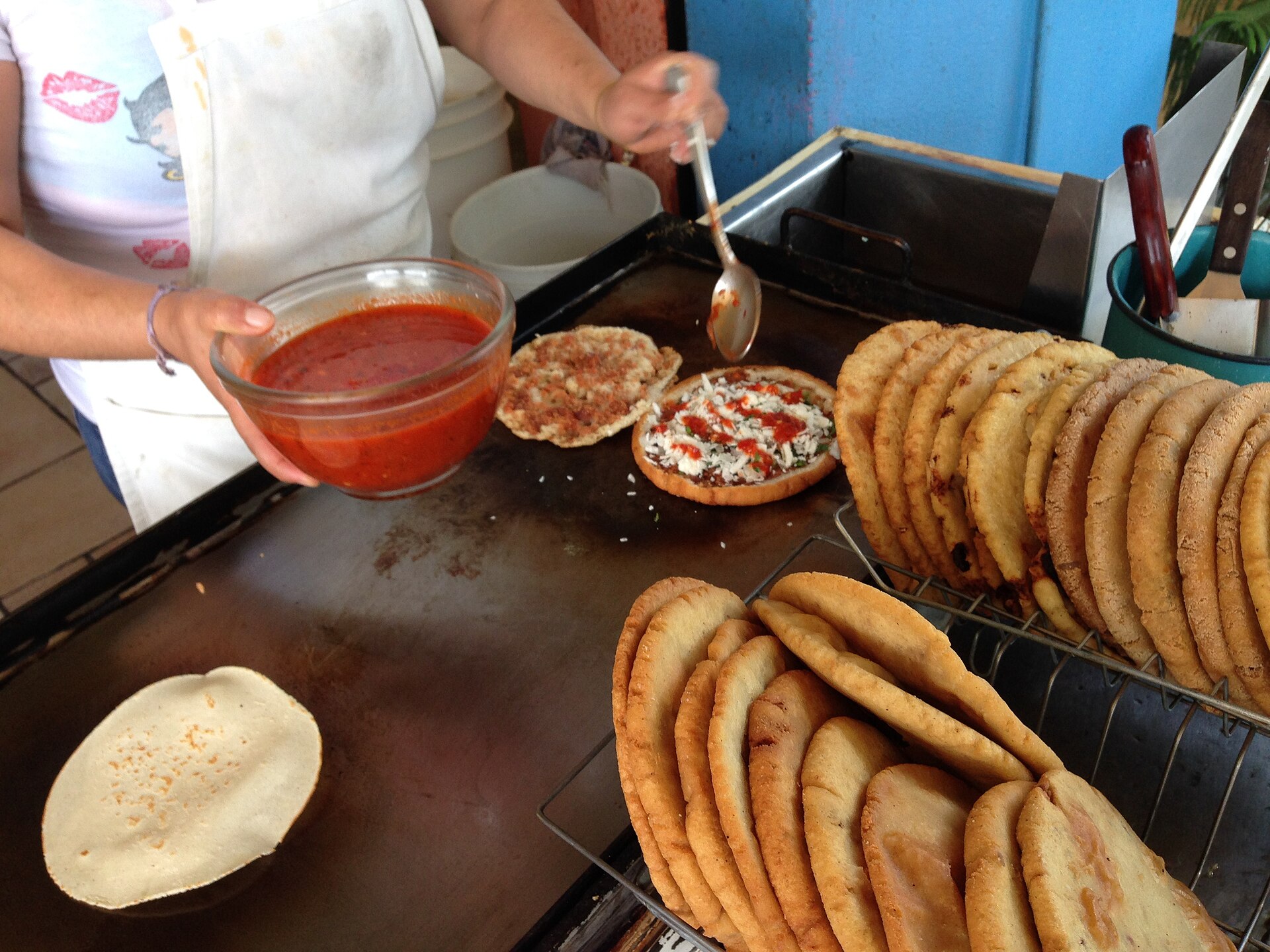 A vendor spoons red salsa onto a split gordita topped with crumbled cheese, beside racks of cooked gorditas on a griddle