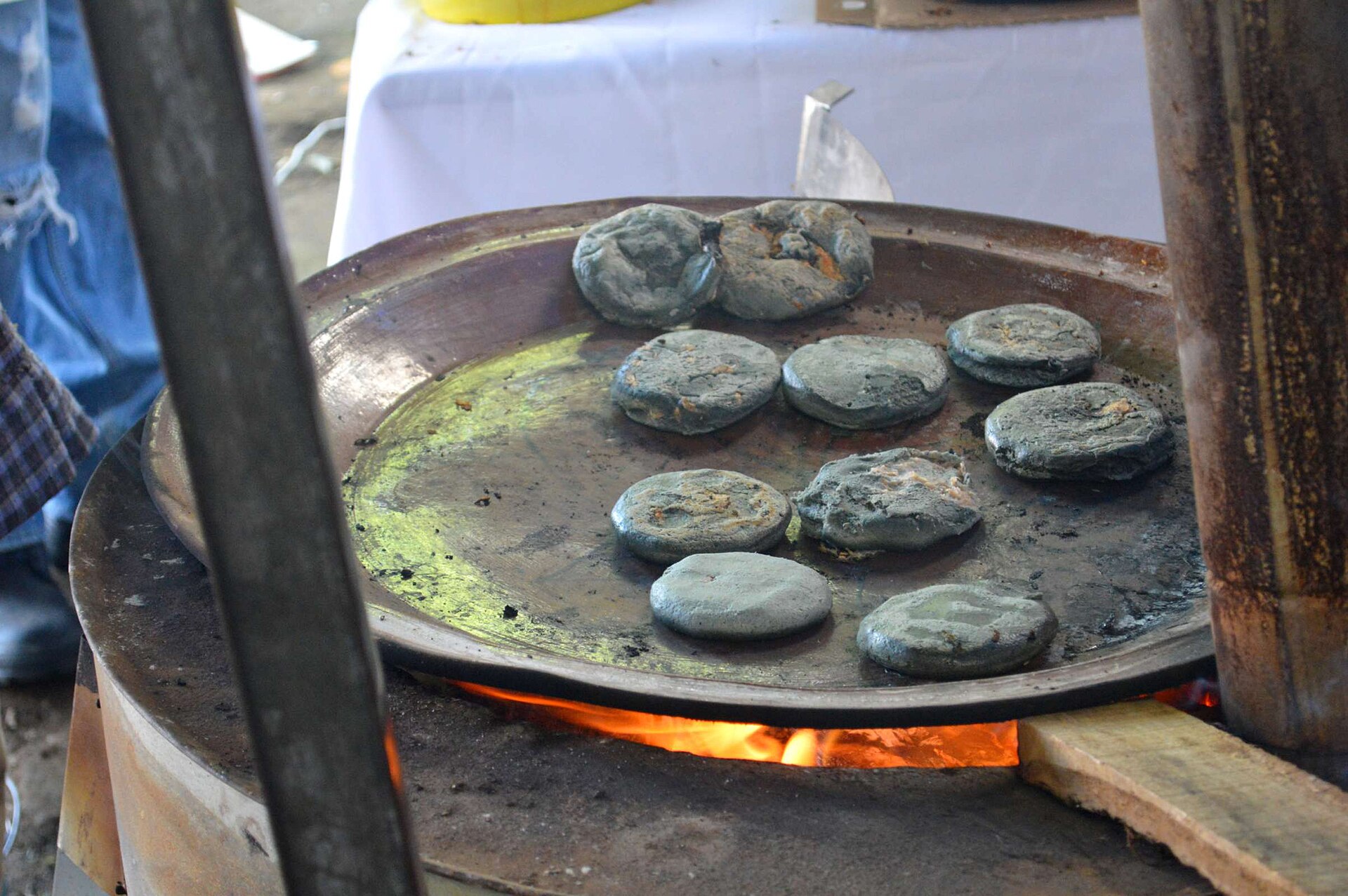 Round blue-grey corn gorditas cooking on a large metal comal over a wood fire