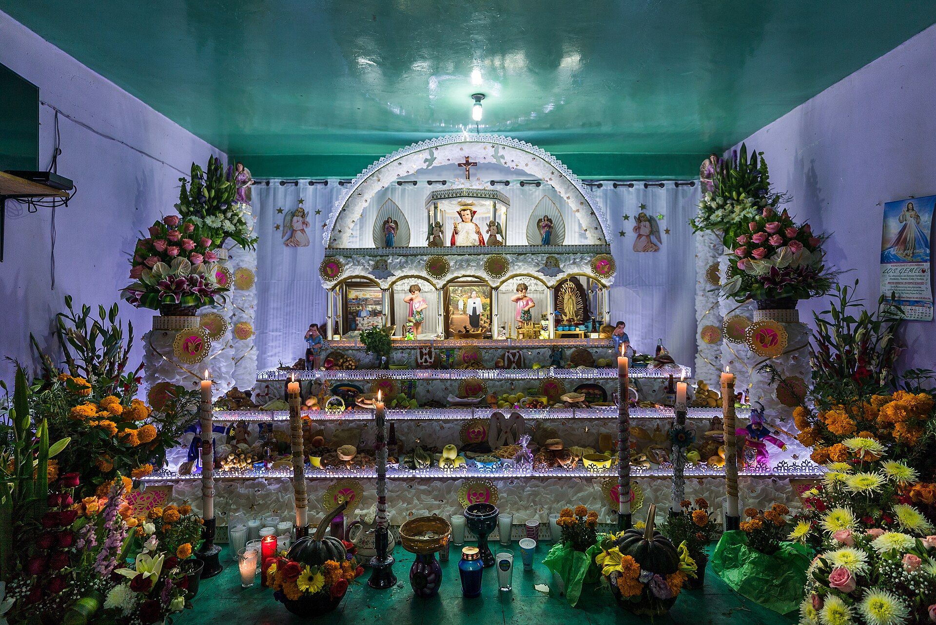 A tiered home altar with candles, food offerings, religious images and vases of orange marigolds and other flowers
