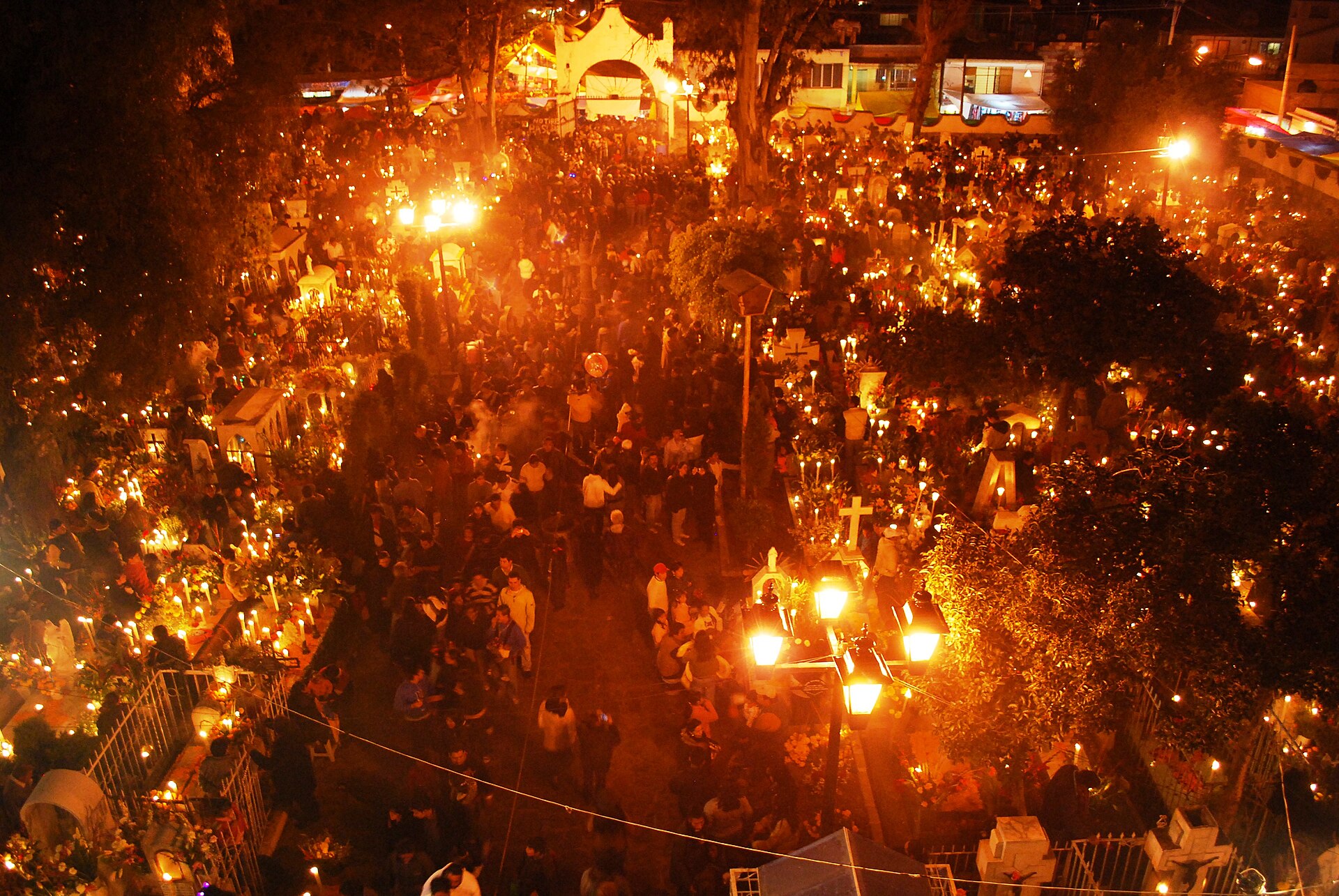A crowded cemetery at night lit by thousands of candles on the graves, seen from above