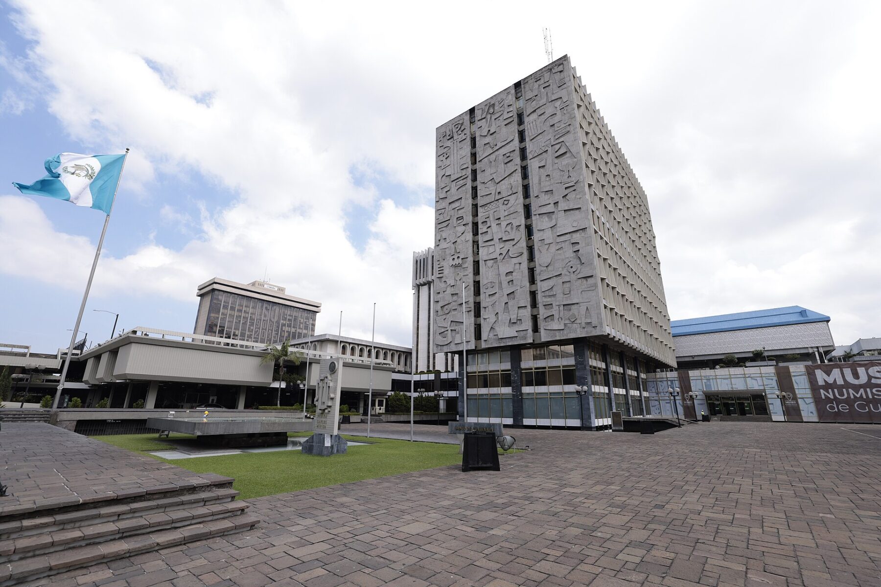 The Bank of Guatemala tower with relief panels, a Guatemalan flag and a paved plaza in front