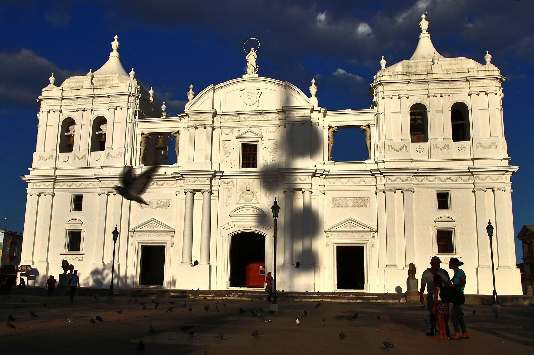 White baroque and neoclassical facade of León Cathedral in Nicaragua with two bell towers and a statue on top, a bird in flight over the plaza at dusk
