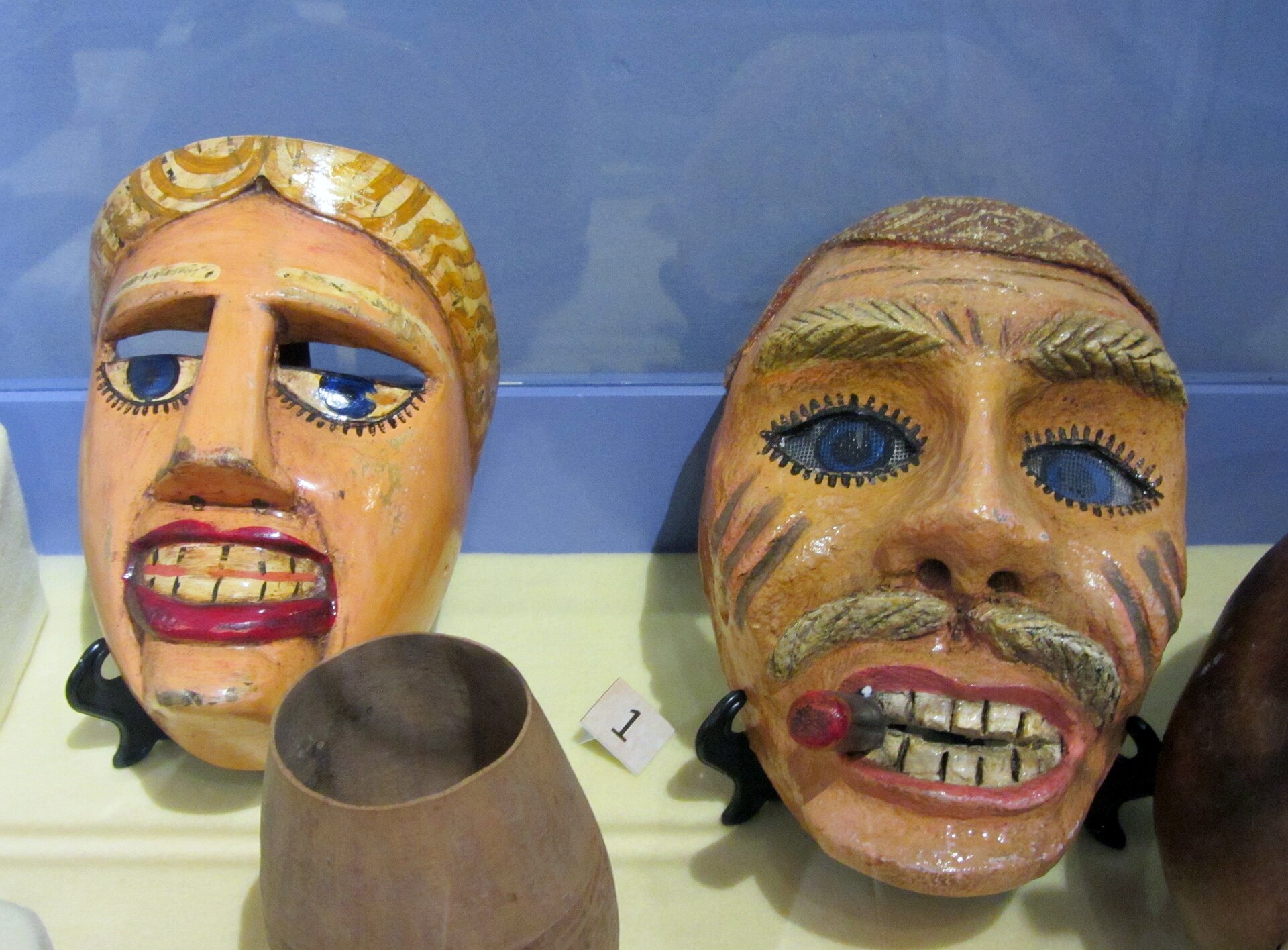 Two painted wooden masks of El Güegüense characters in a museum display case, one with a yellow headcloth and one with a moustache and a cigar