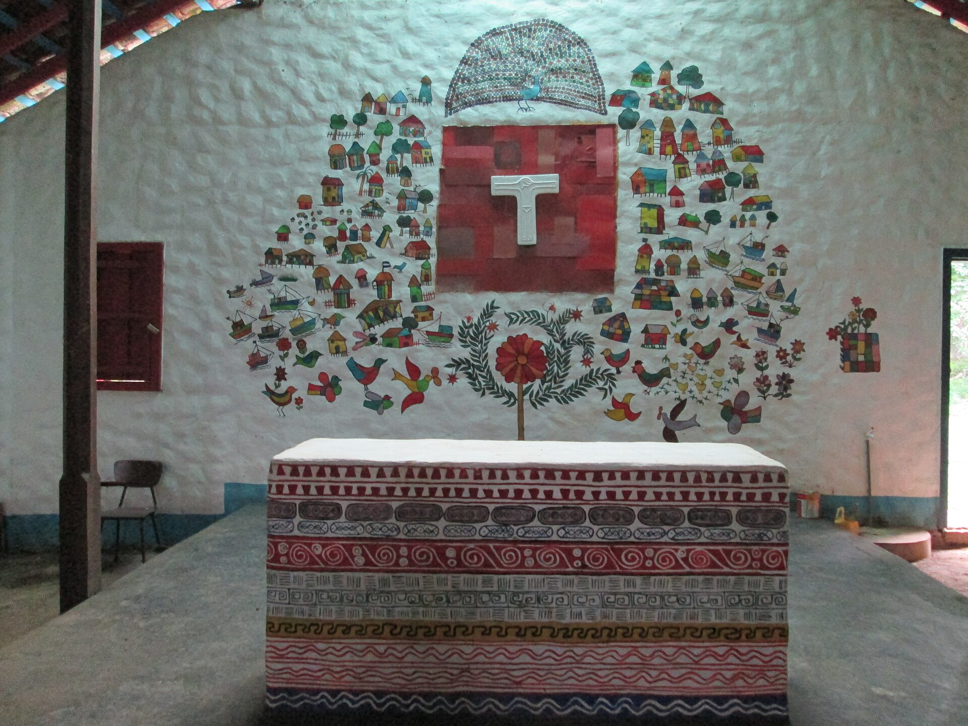 Whitewashed church wall in Solentiname with a painted folk mural of houses, boats, birds and a flower around a white cross, and a painted altar block in front