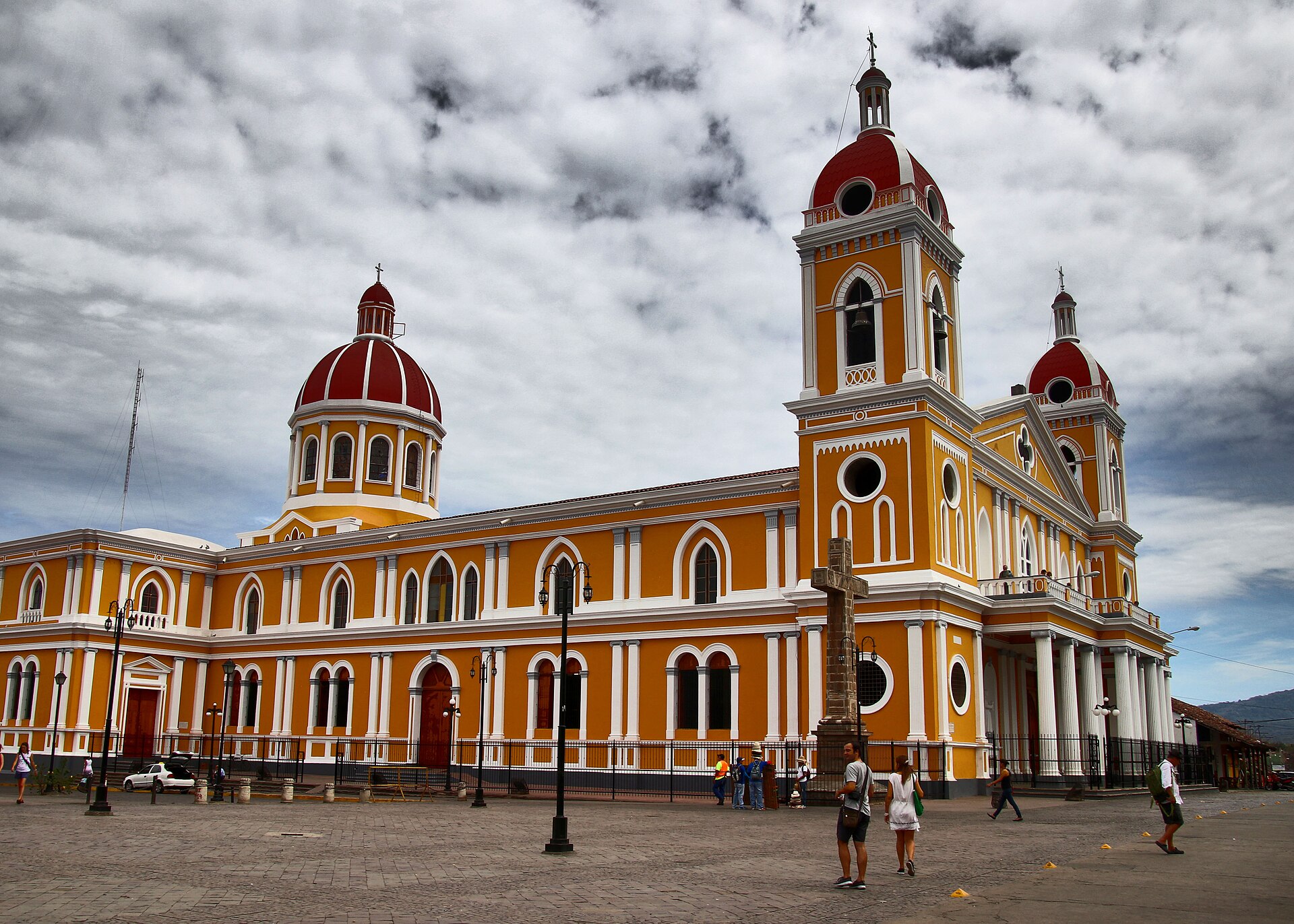 Yellow and white cathedral of Granada, Nicaragua, with red domes and a bell tower under a cloudy sky, people crossing the square in front