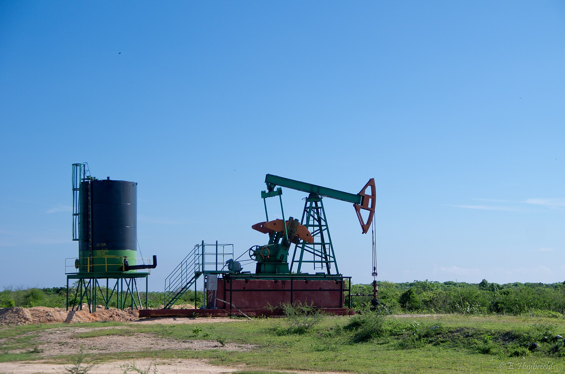 A green and orange oil pumpjack beside a black storage tank on scrubland in Cuba under a clear blue sky