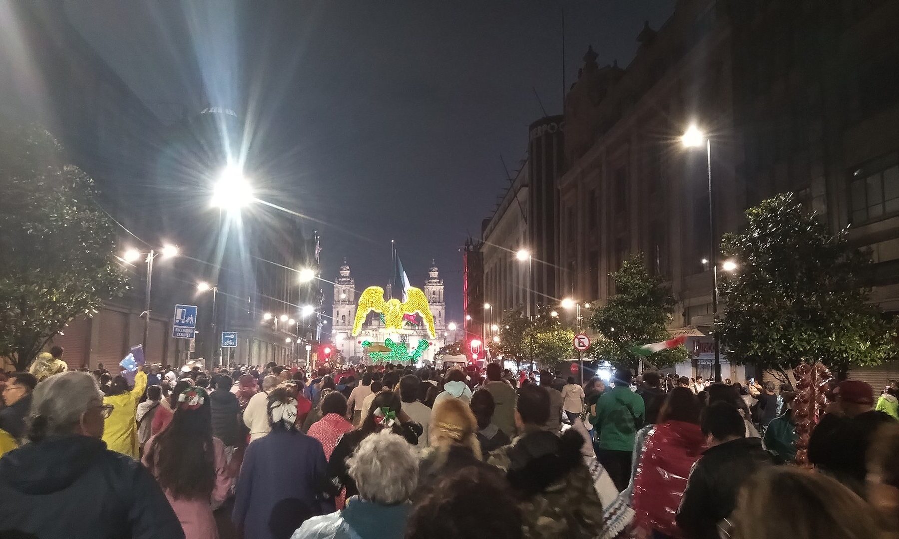 Crowds walking at night along a Mexico City street towards the Zócalo, where a lit-up eagle decoration and the cathedral towers glow at the end of the street