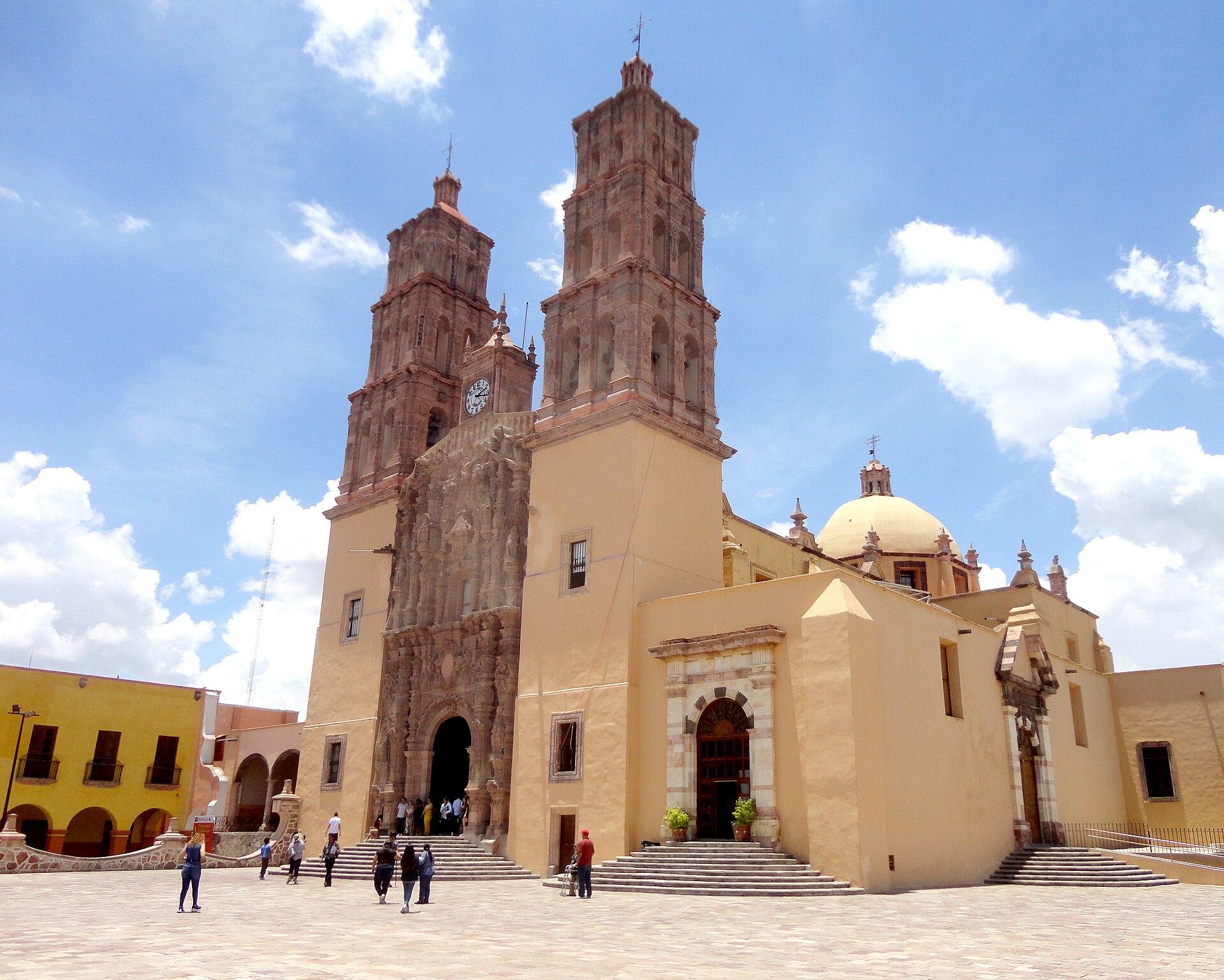 A church with two reddish stone bell towers, ochre walls and a dome on a wide paved square under a blue sky with clouds