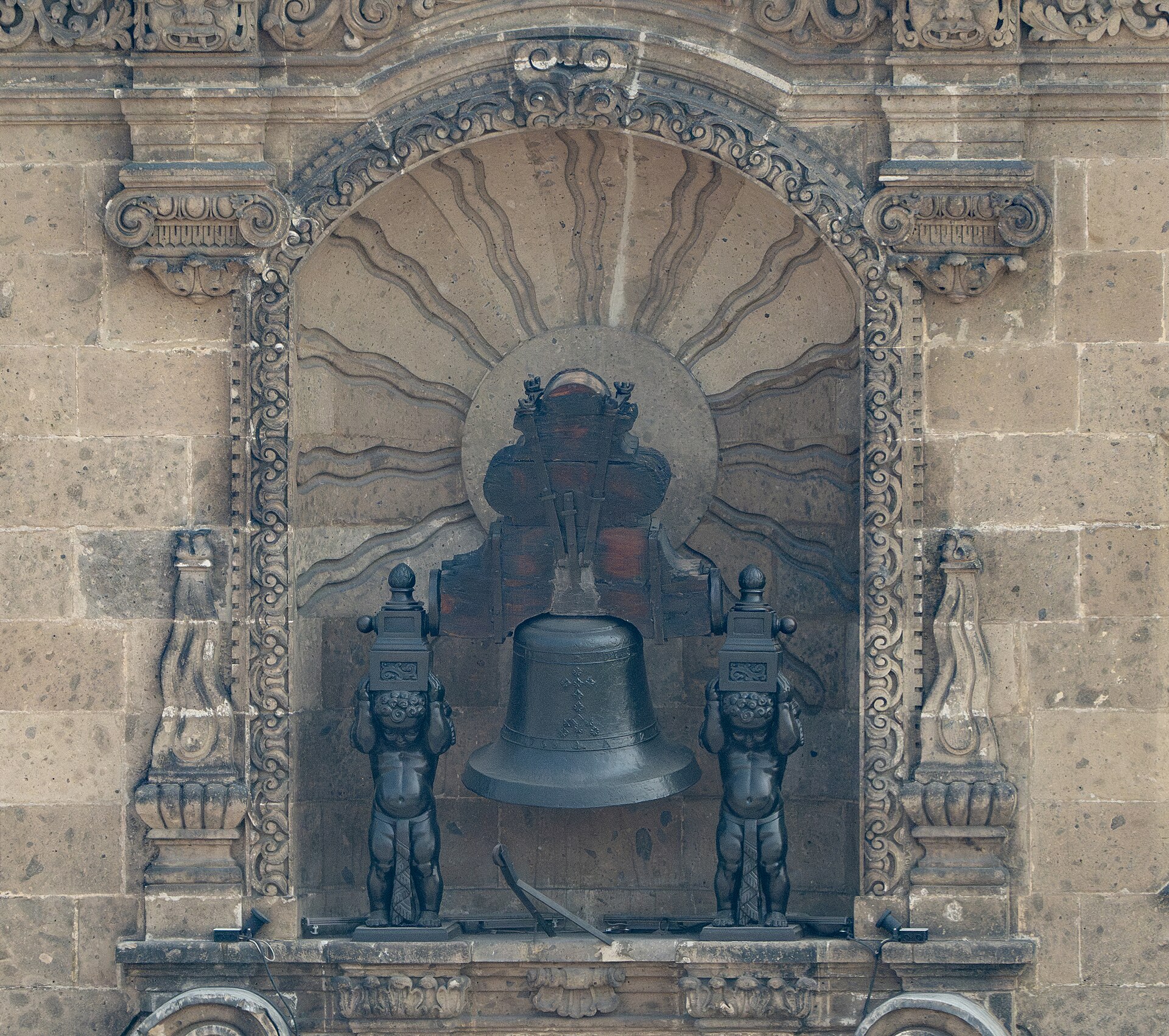 A dark bronze bell hanging in a carved stone niche with a sunburst relief, flanked by two small dark statues