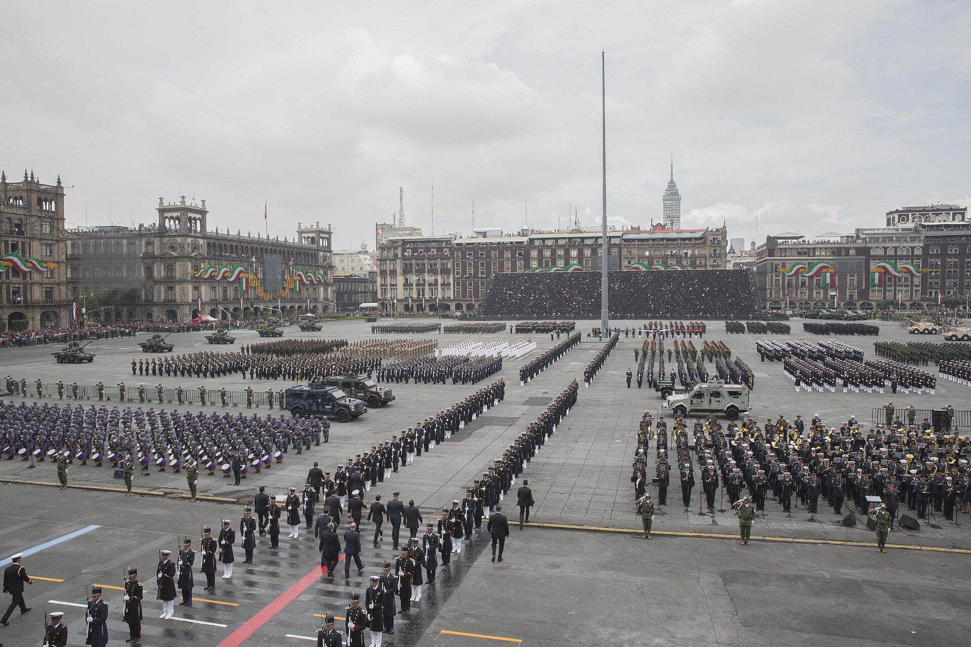 Rows of uniformed troops, military bands and armoured vehicles lined up across Mexico City's Zócalo under a grey sky, with a giant flagpole in the centre