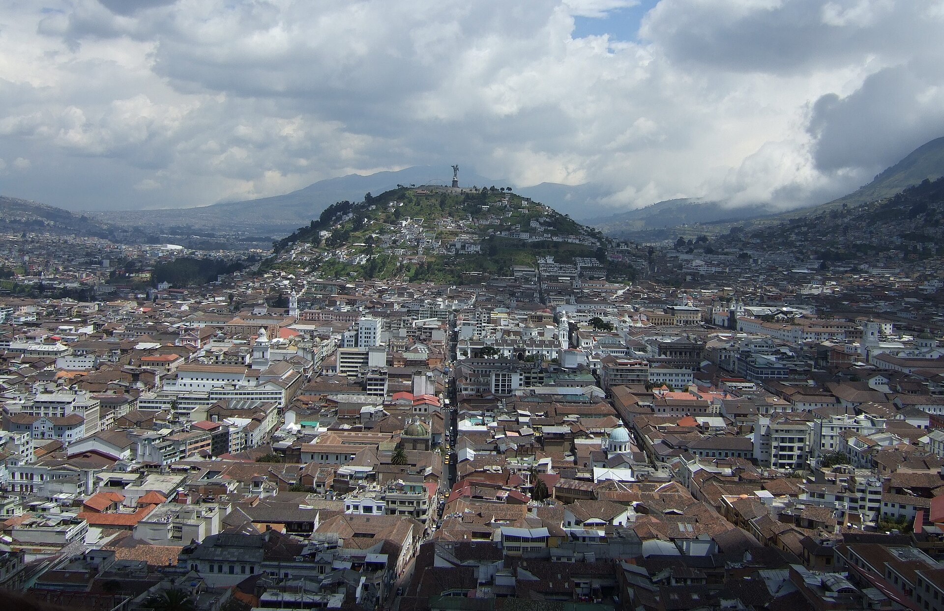 Dense rooftops of central Quito stretching towards a green hill topped by a winged Virgin statue under cloudy skies