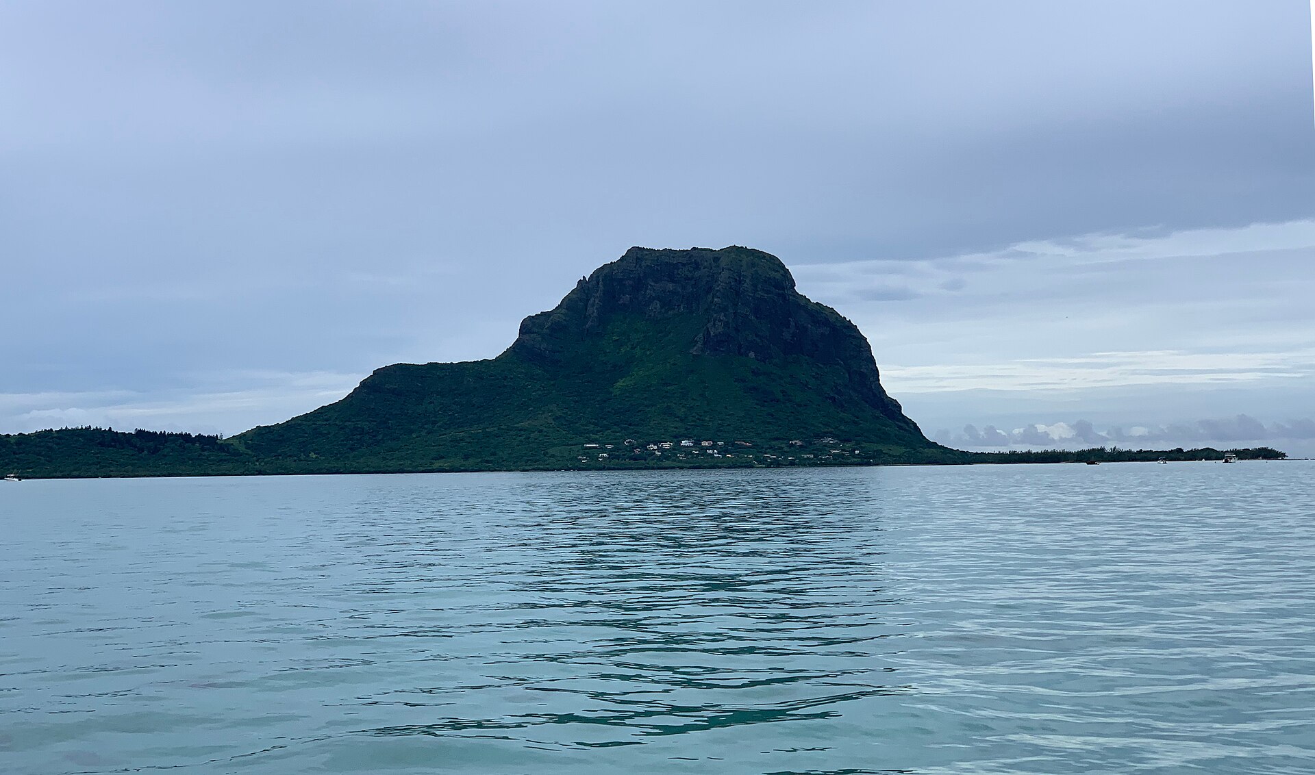 Green basalt mountain of Le Morne Brabant rising from calm turquoise water under a grey sky