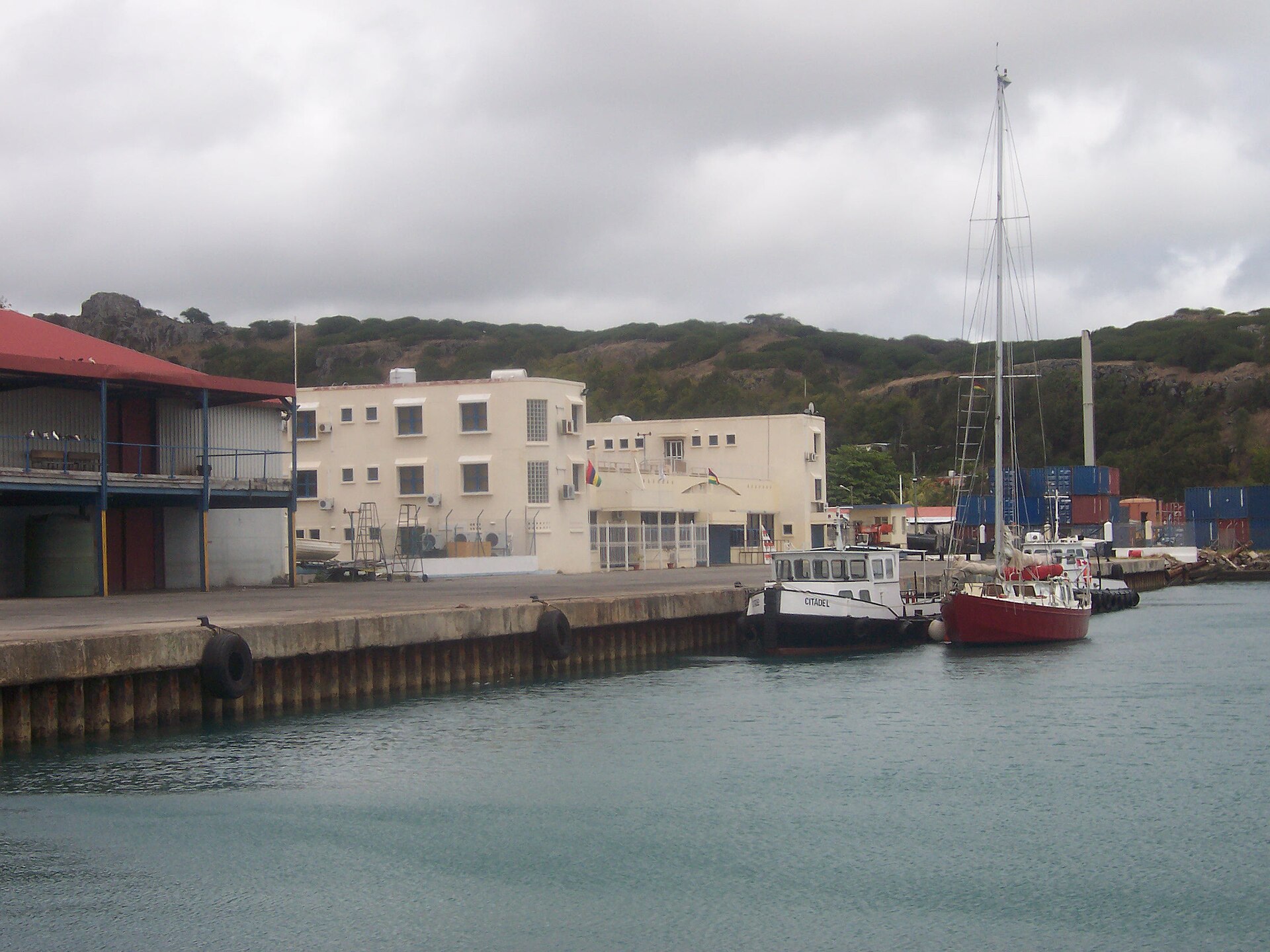 A concrete quay with a tugboat and a red sailing yacht, a cream coloured two-storey building and stacked containers behind, under a cloudy sky