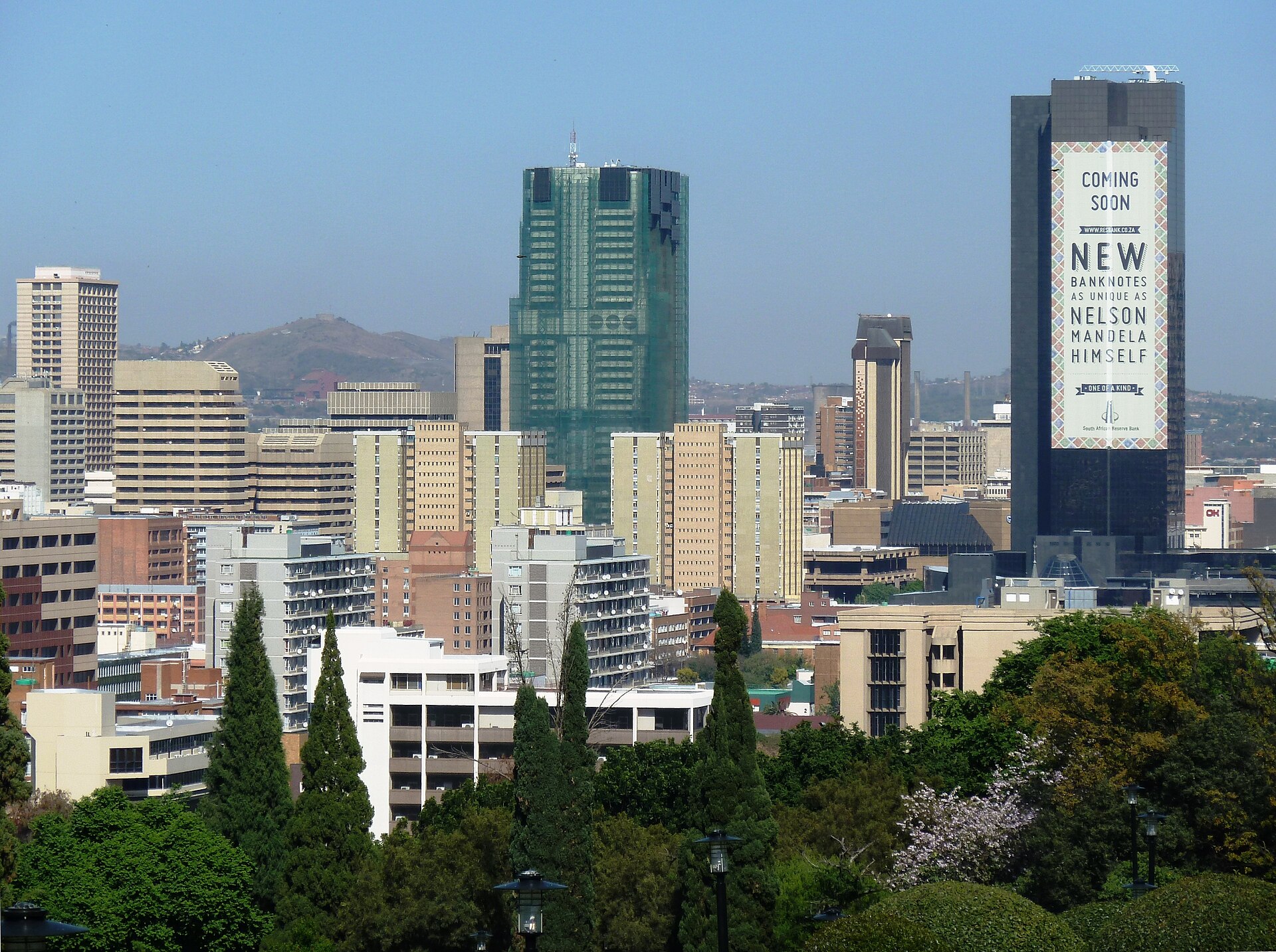 Pretoria's city centre towers behind trees, with a dark high-rise on the right carrying a large billboard for new banknotes