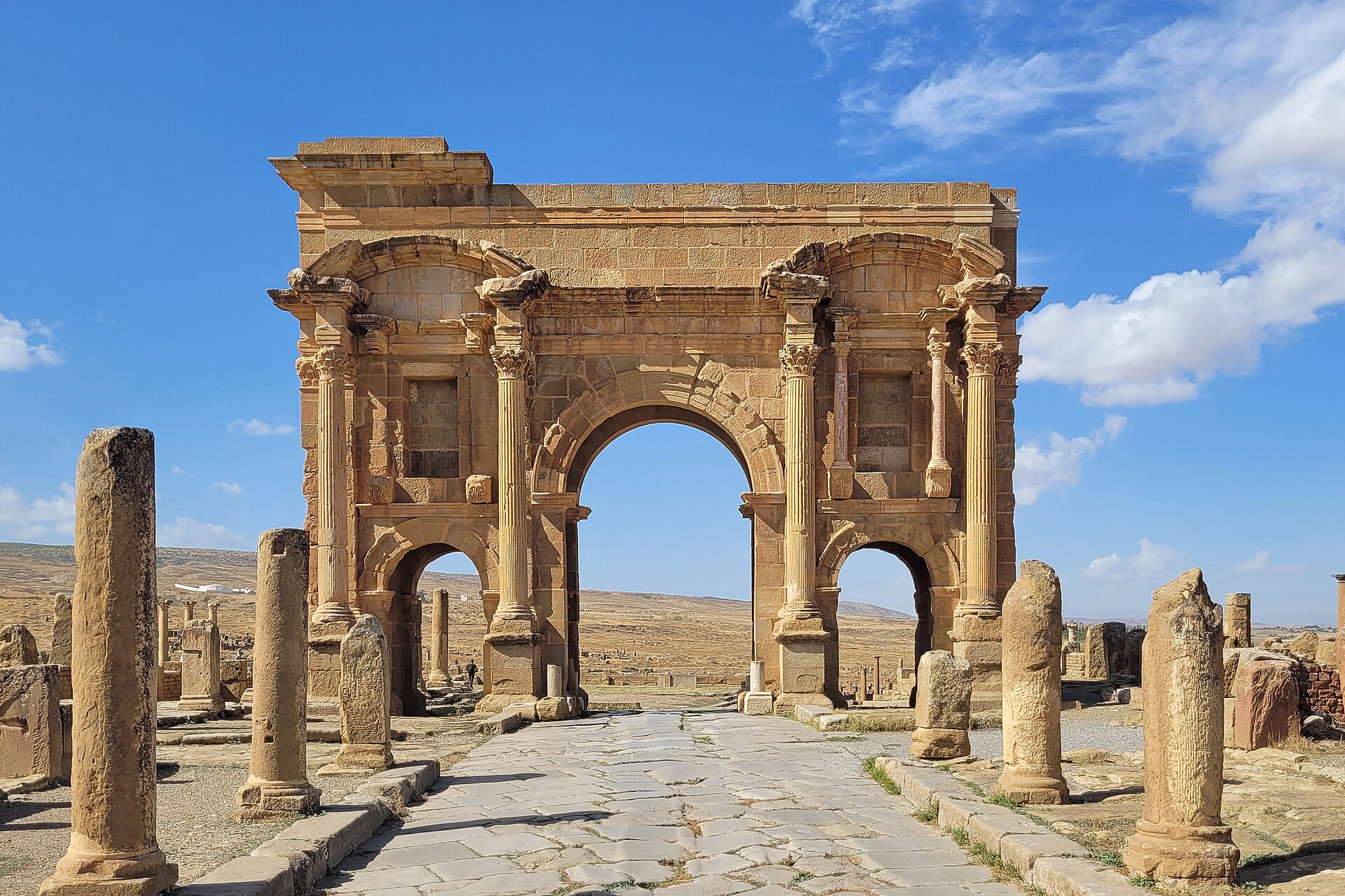 The Arch of Trajan at Timgad with weathered columns, a paved Roman street and a blue sky