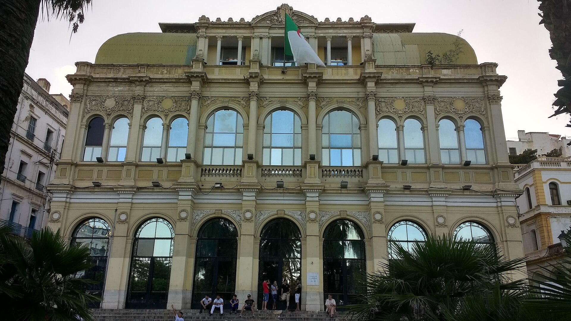 Ornate cream facade of the Algerian National Theatre with arched windows and an Algerian flag, seen from the steps