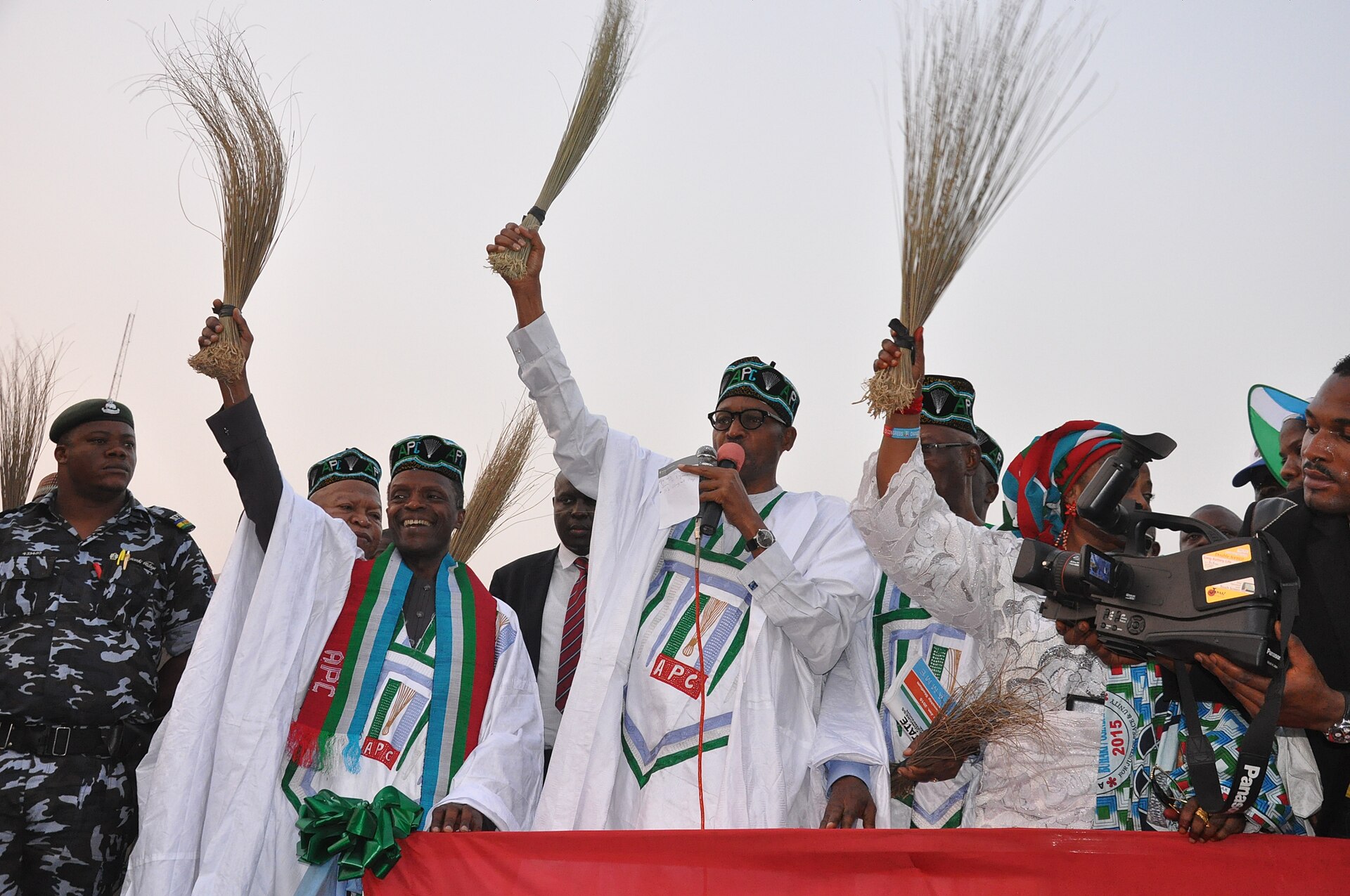 Men in white robes and green caps marked APC raise brooms on a stage while a police officer and a cameraman stand nearby