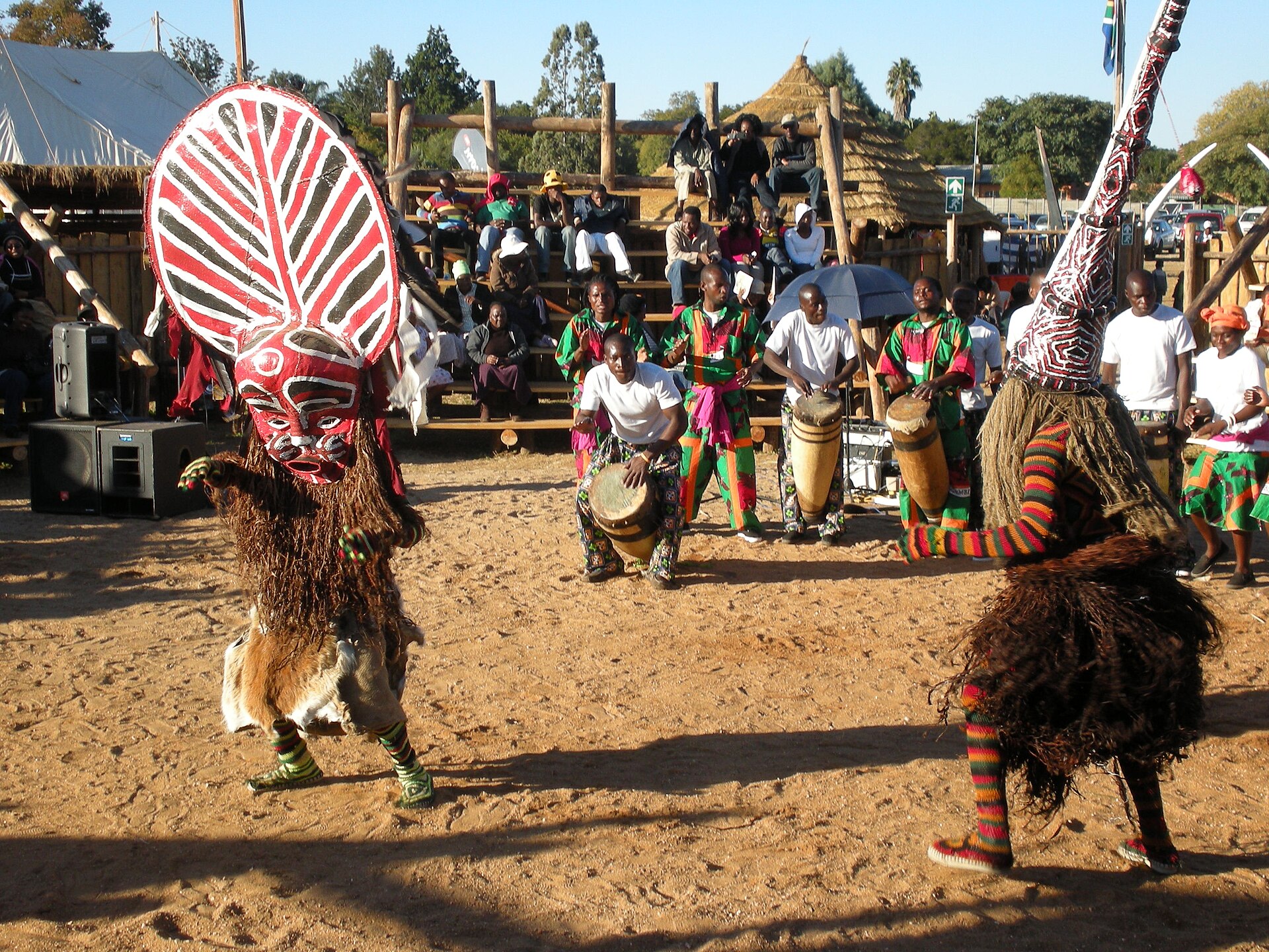 Two masked Makishi dancers in raffia costumes, one with a red and white striped mask, perform on sand while drummers in green and pink sit behind
