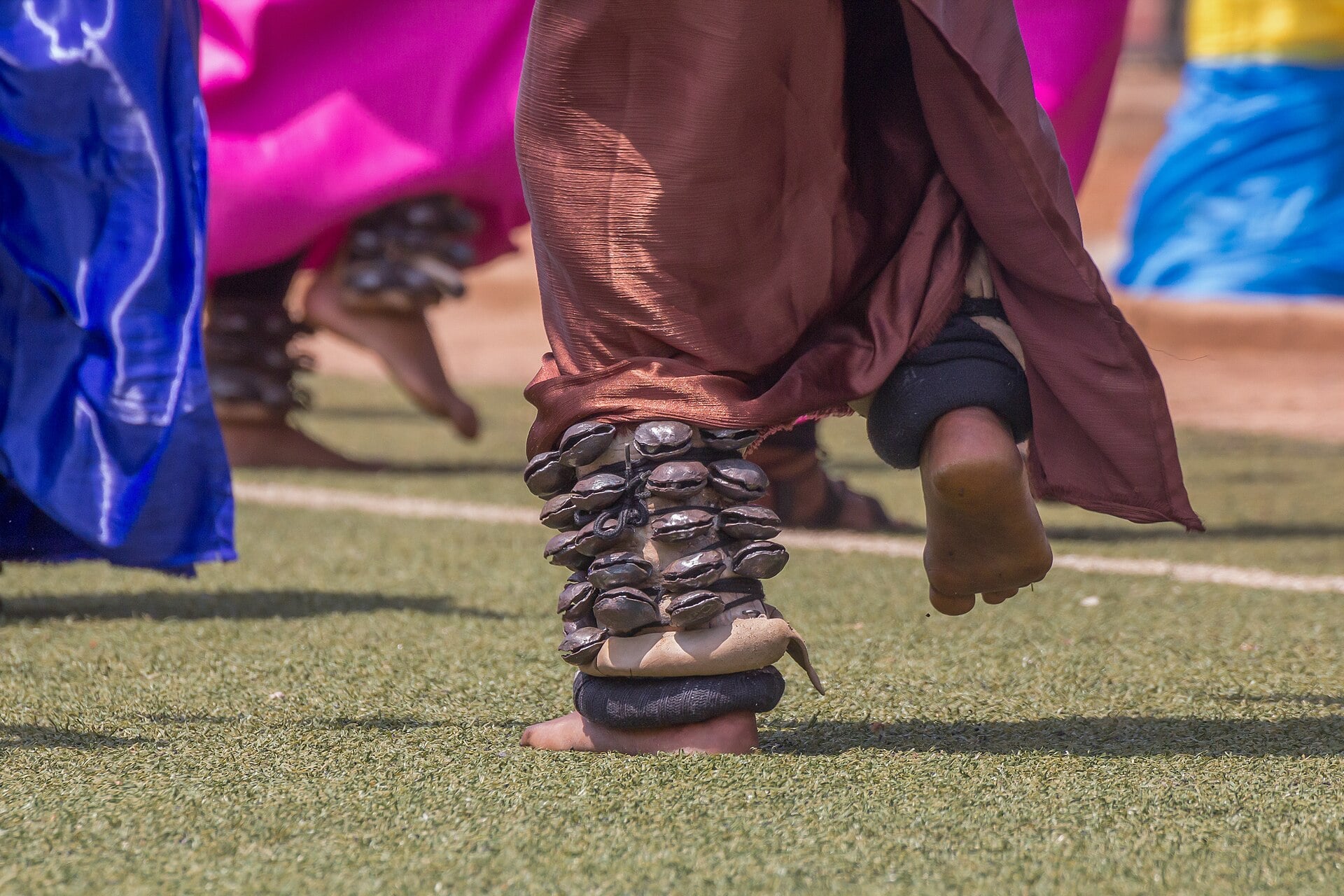 Close view of an Intore dancer's feet with ankle rattles, among dancers in colourful wraps on a grass field