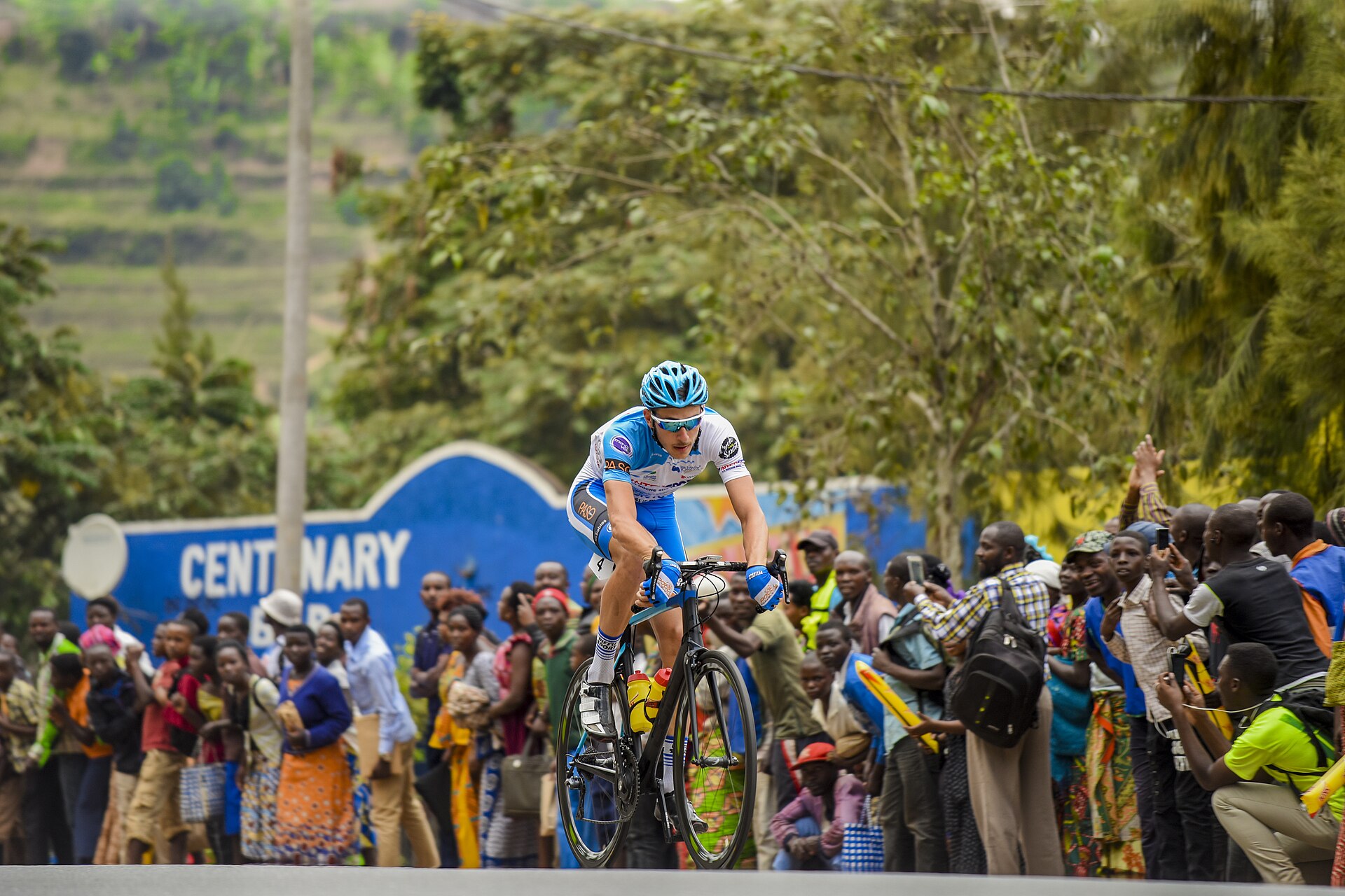 Cyclist in a blue and white kit riding uphill past a crowd of roadside spectators in Rwanda