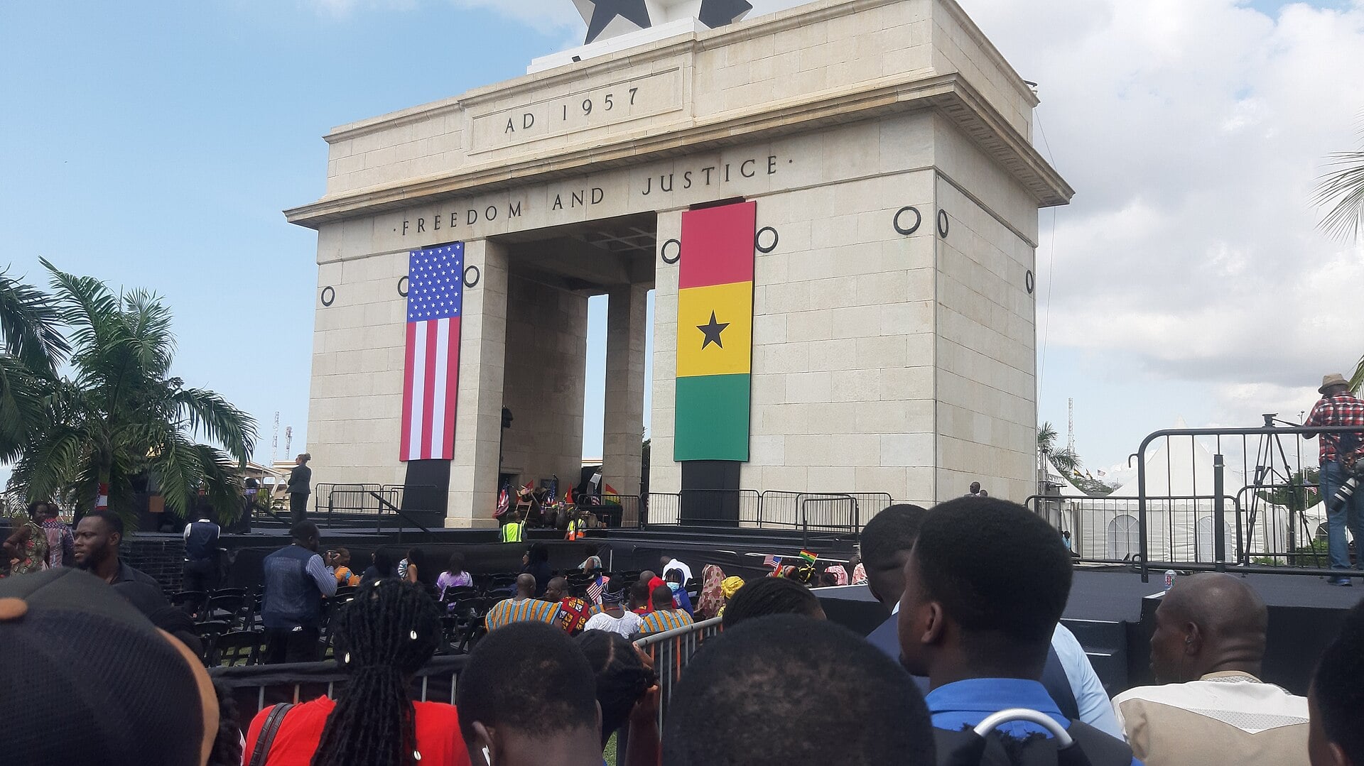 The Independence Arch at Black Star Square in Accra, hung with a US flag and a Ghanaian flag, with a crowd and palm trees in front