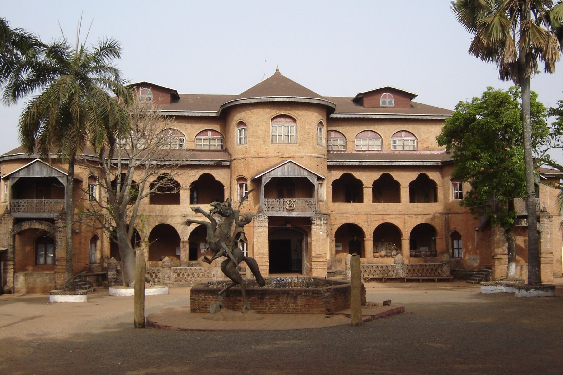 Two-storey red-brown brick palace in Foumban with arched verandas, a bronze horse statue on a fountain in the courtyard and palm trees