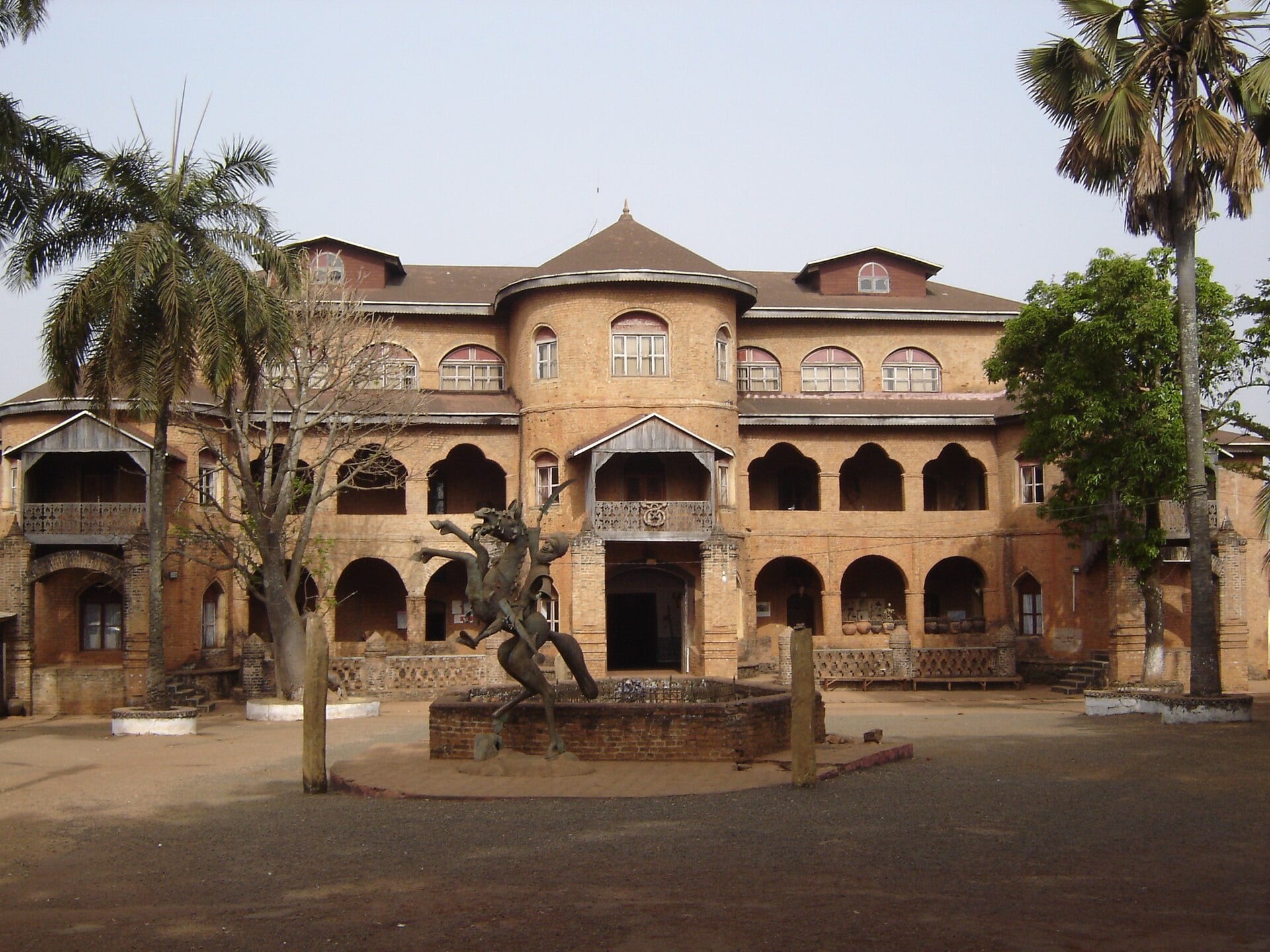 Two-storey red-brown brick palace in Foumban with arched verandas, a bronze horse statue on a fountain in the courtyard and palm trees