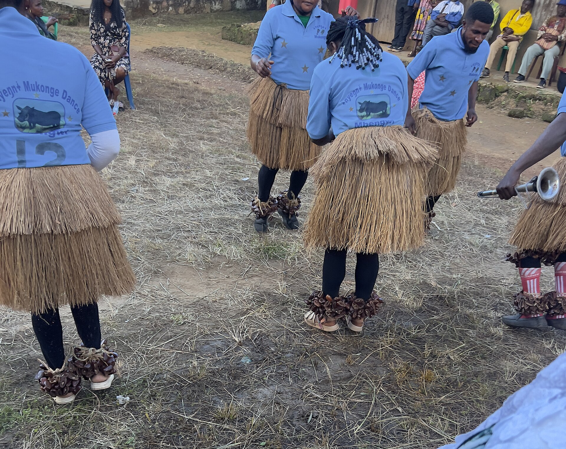 Dancers in blue shirts and raffia skirts with bead-and-seed ankle rattles dance on dry grass while a man plays a horn and spectators sit behind