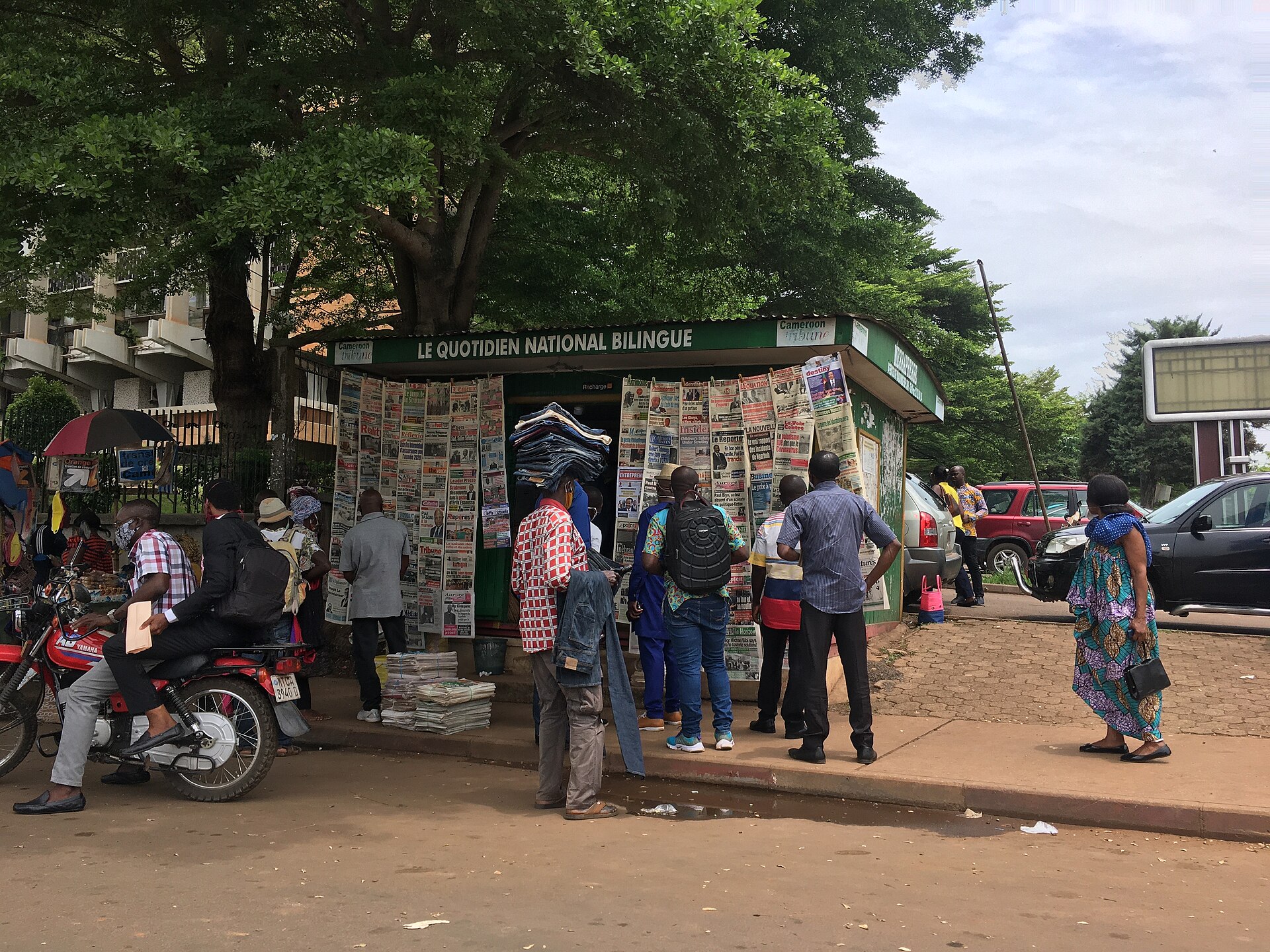 People reading newspapers pinned to a green kiosk on a Yaoundé street with motorbikes and trees around it