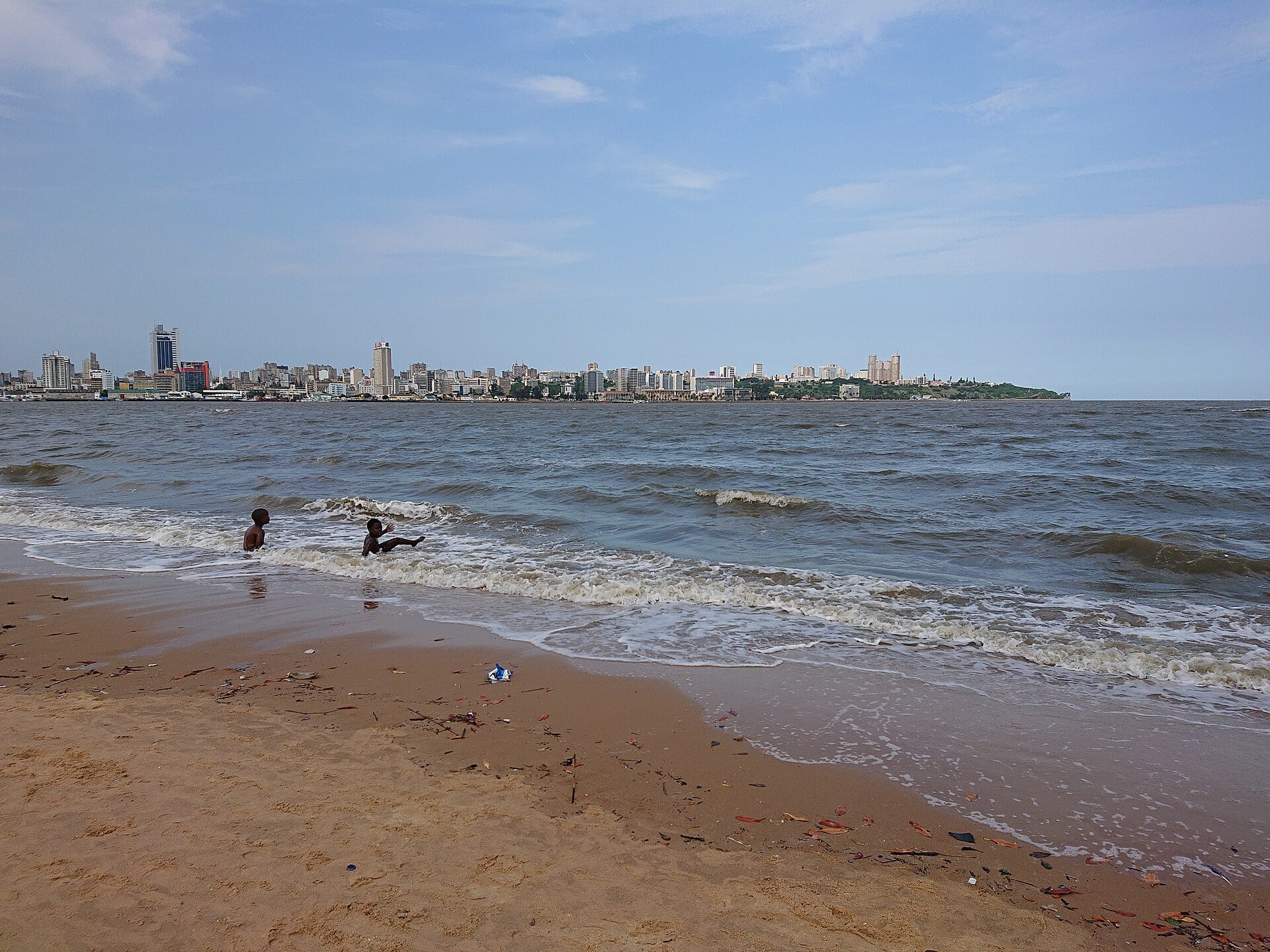The skyline of Maputo with office and apartment towers seen across the waters of Maputo Bay, with two children playing in the surf on a sandy beach in the foreground