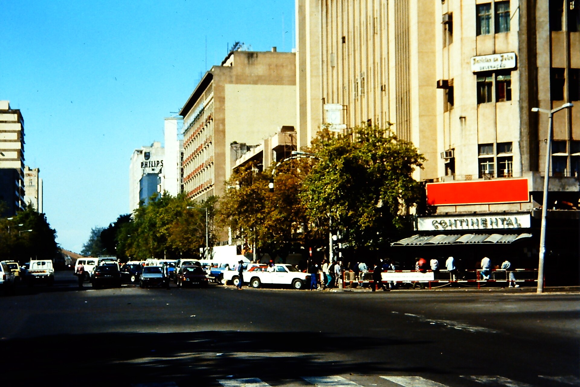 A wide street in downtown Maputo lined with multi-storey office and commercial buildings, trees, parked cars and pedestrians under a clear blue sky