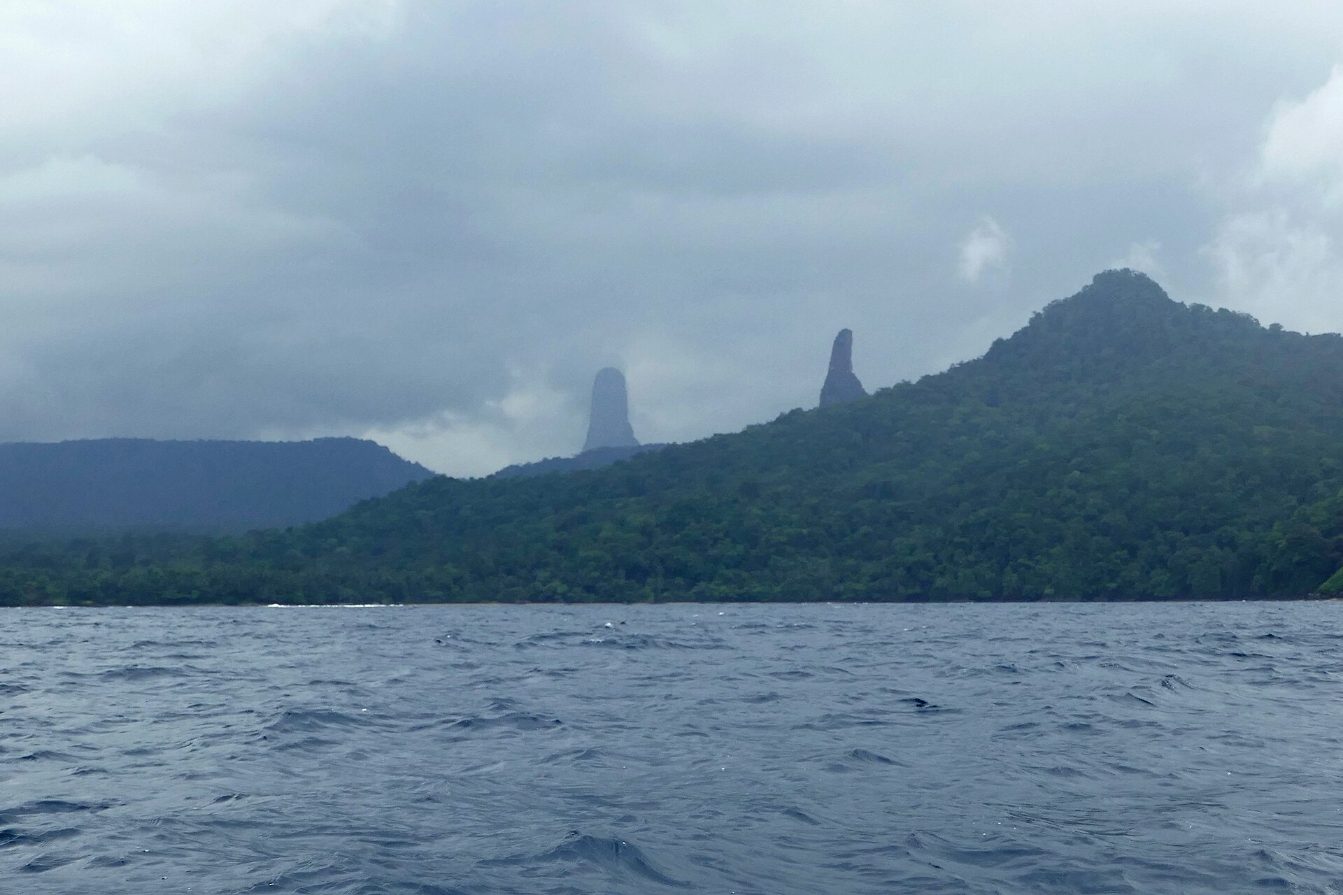 Two volcanic rock spires, Cão Grande and Cão Pequeno, rise above forested hills on São Tomé island, seen from the sea under heavy cloud