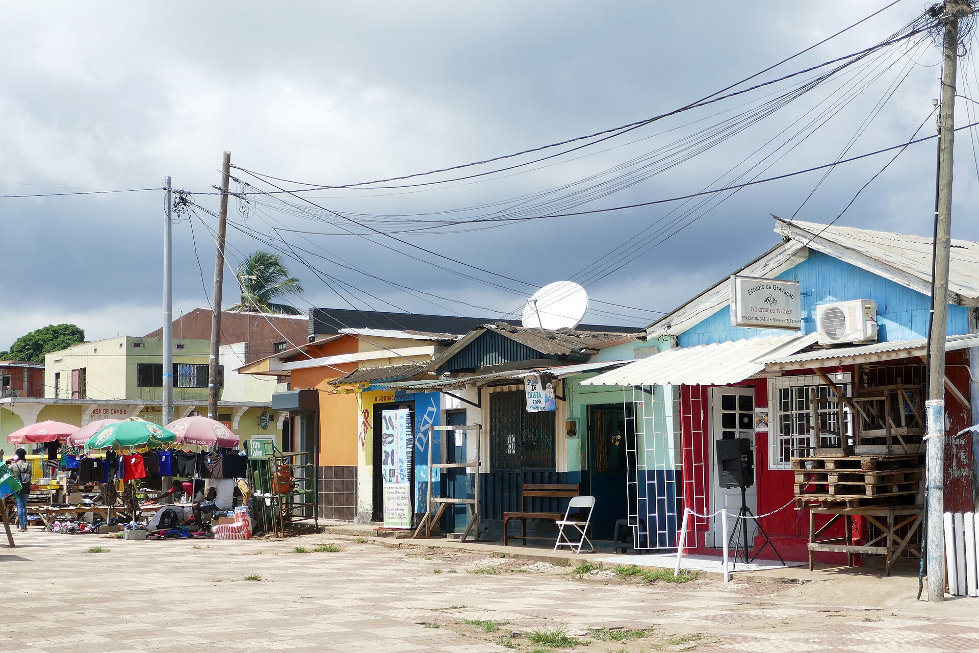 Street scene at the market square of São Tomé with market stalls under umbrellas, painted wooden kiosks, a satellite dish and overhead cables
