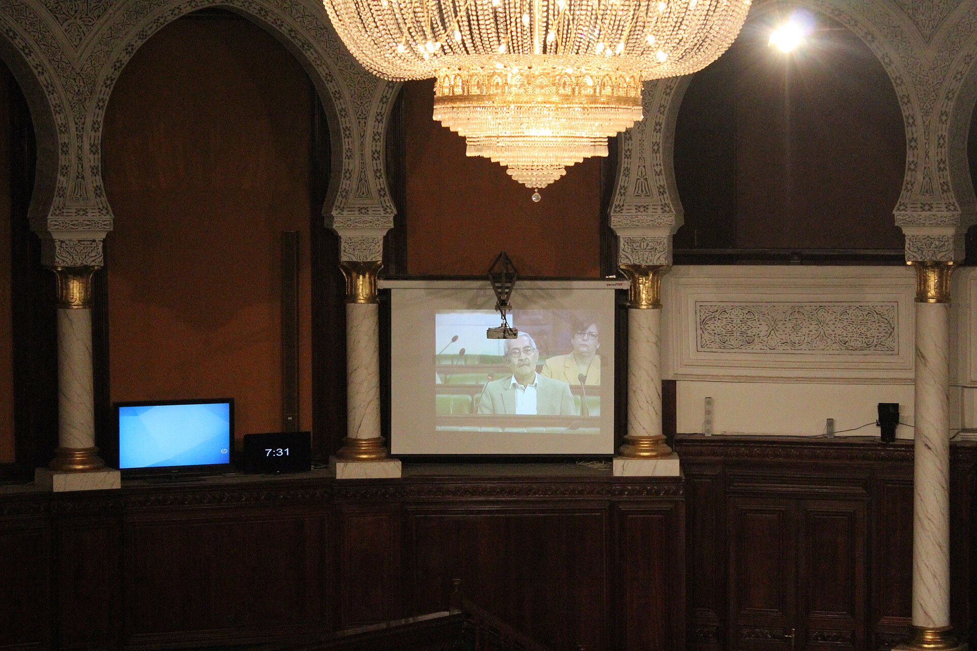 Ornate hall with horseshoe arches, marble columns and a chandelier, with a projection screen showing a speaker in a suit