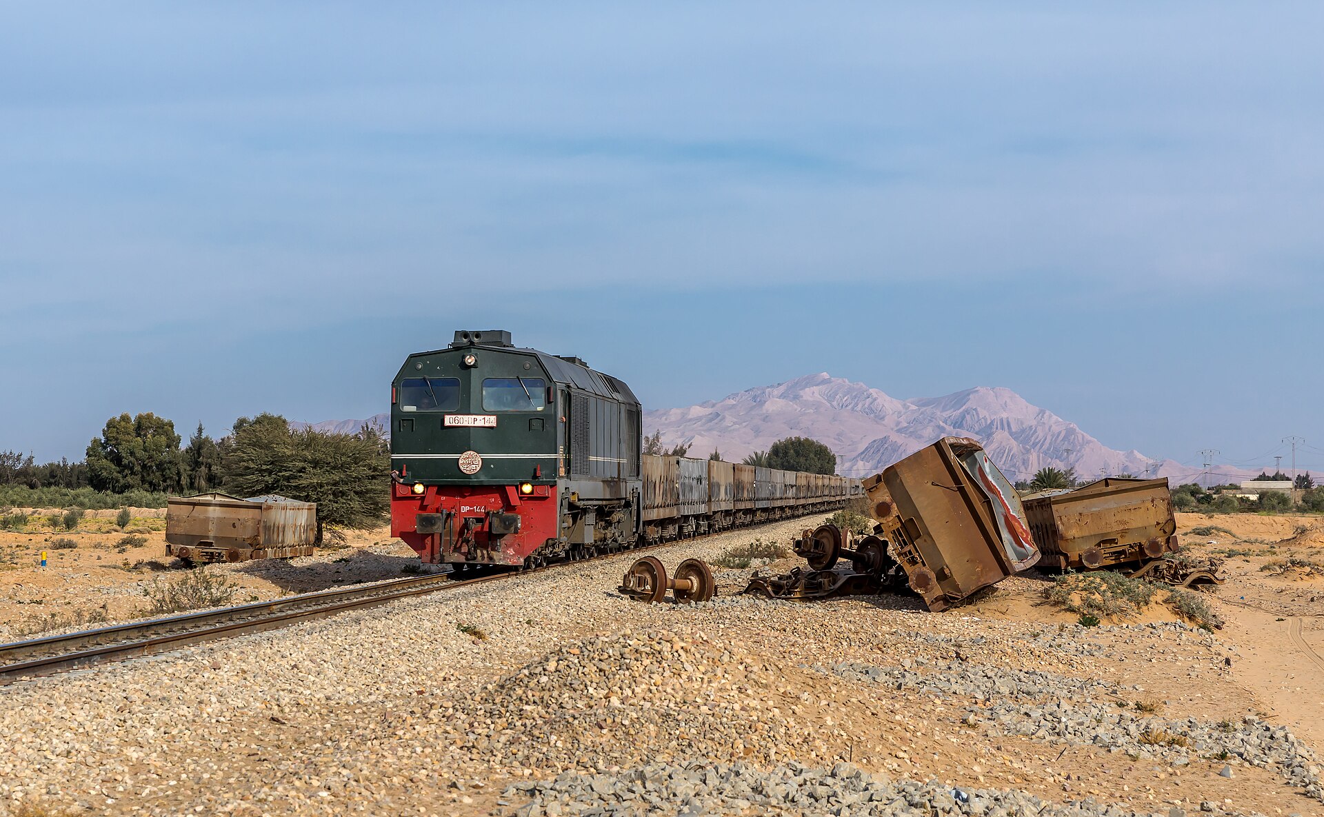 A green and red diesel locomotive pulling a long train of open wagons through desert scrub with bare mountains behind and a derailed wagon beside the track
