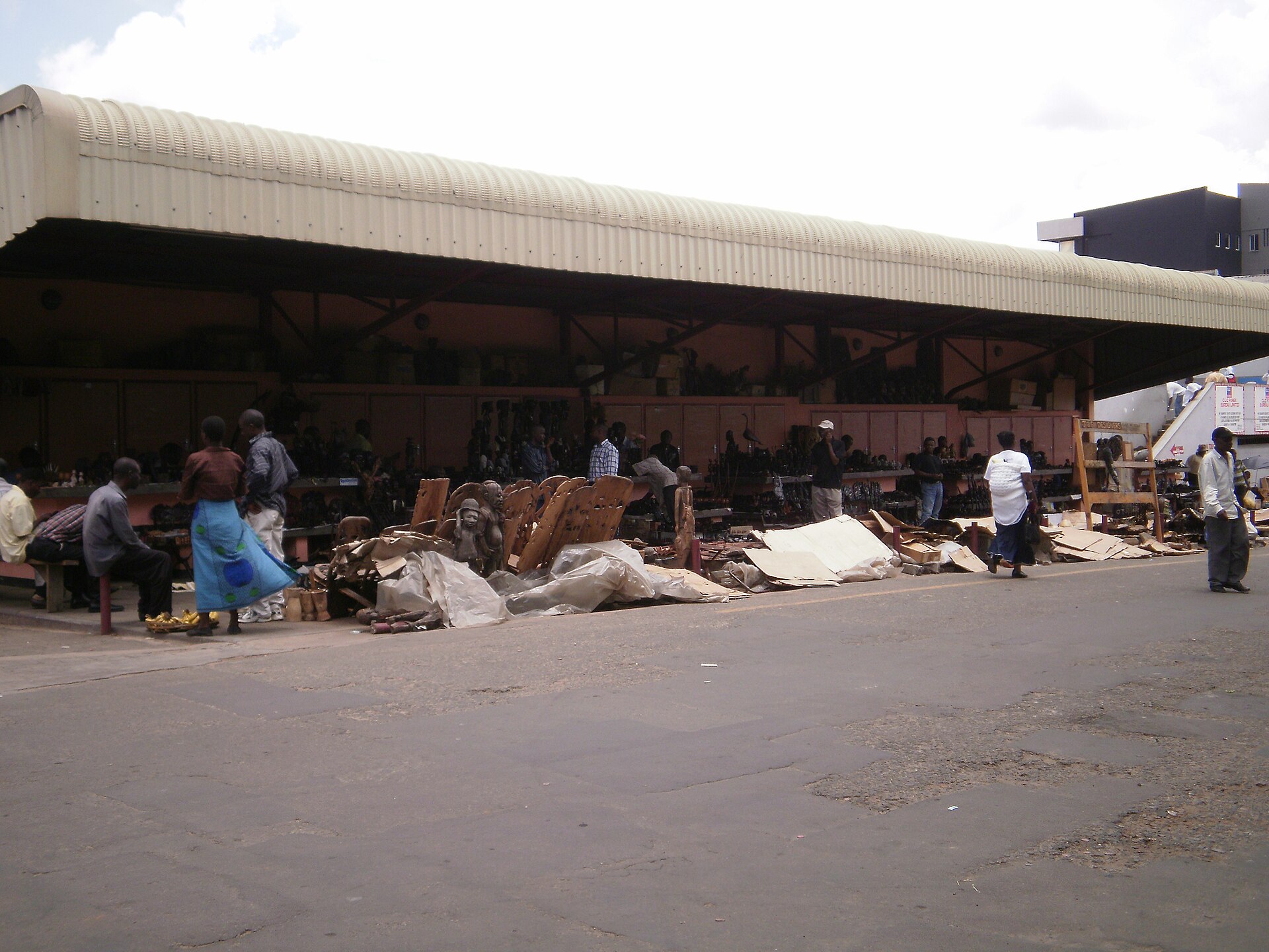 People standing and sitting outside a covered craft market with carved wooden items in Blantyre, Malawi