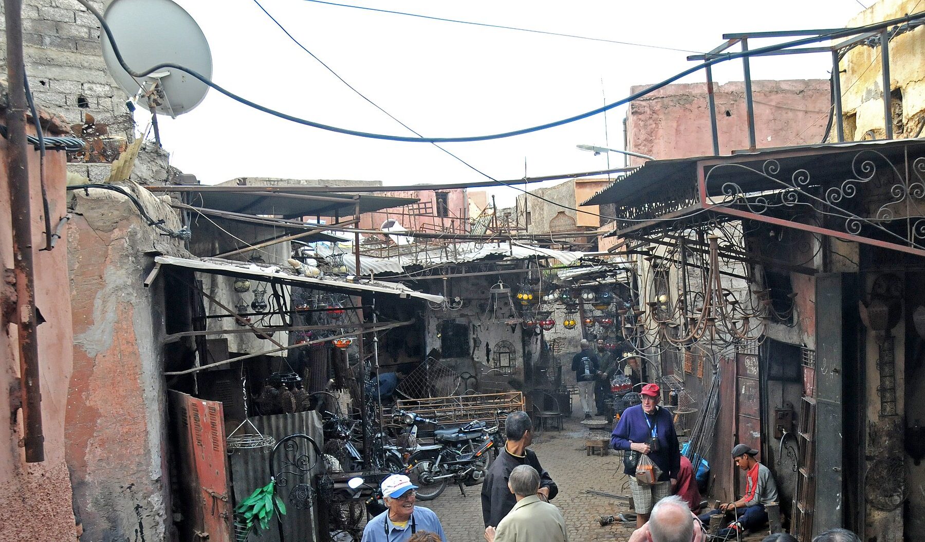 A narrow lane in the Marrakech medina lined with metalworkers' workshops, lanterns hanging under tin awnings and visitors standing among shopkeepers